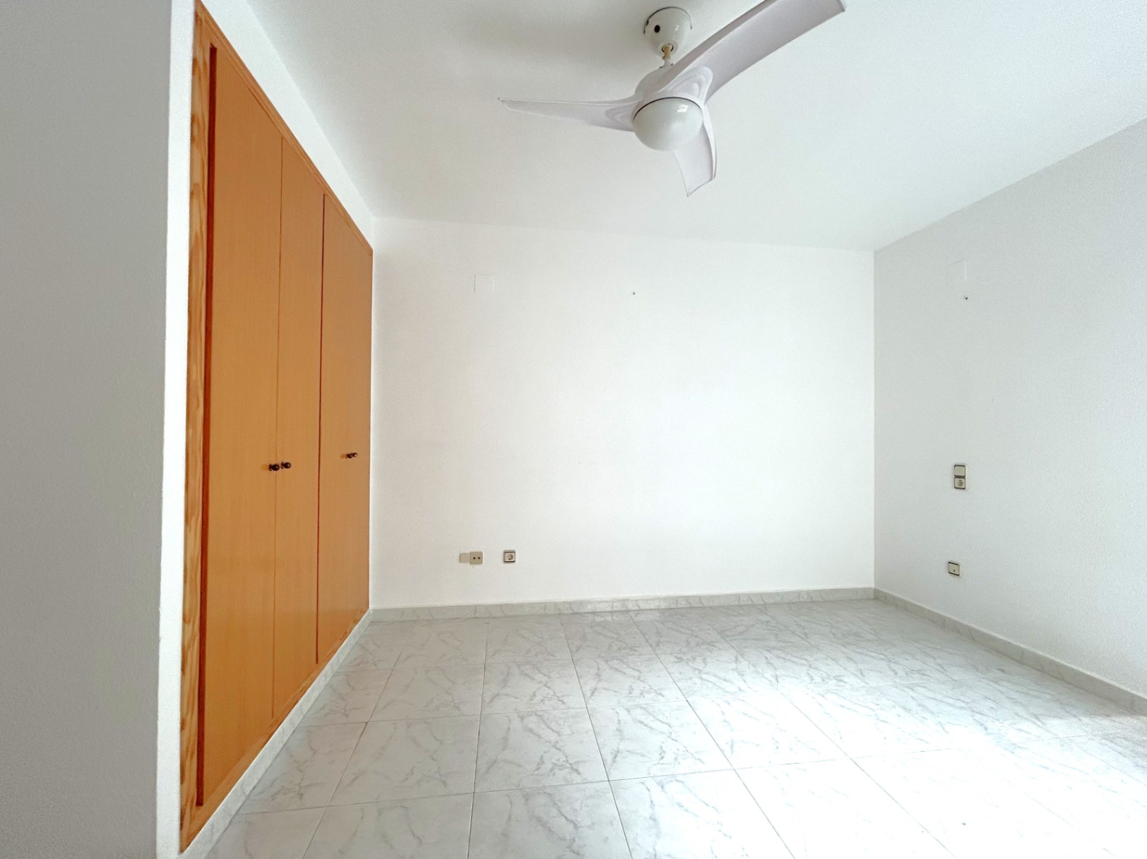 Chambre lumineuse avec grand placard intégré en bois, sol en grès effet marbre et ventilateur de plafond blanc.
