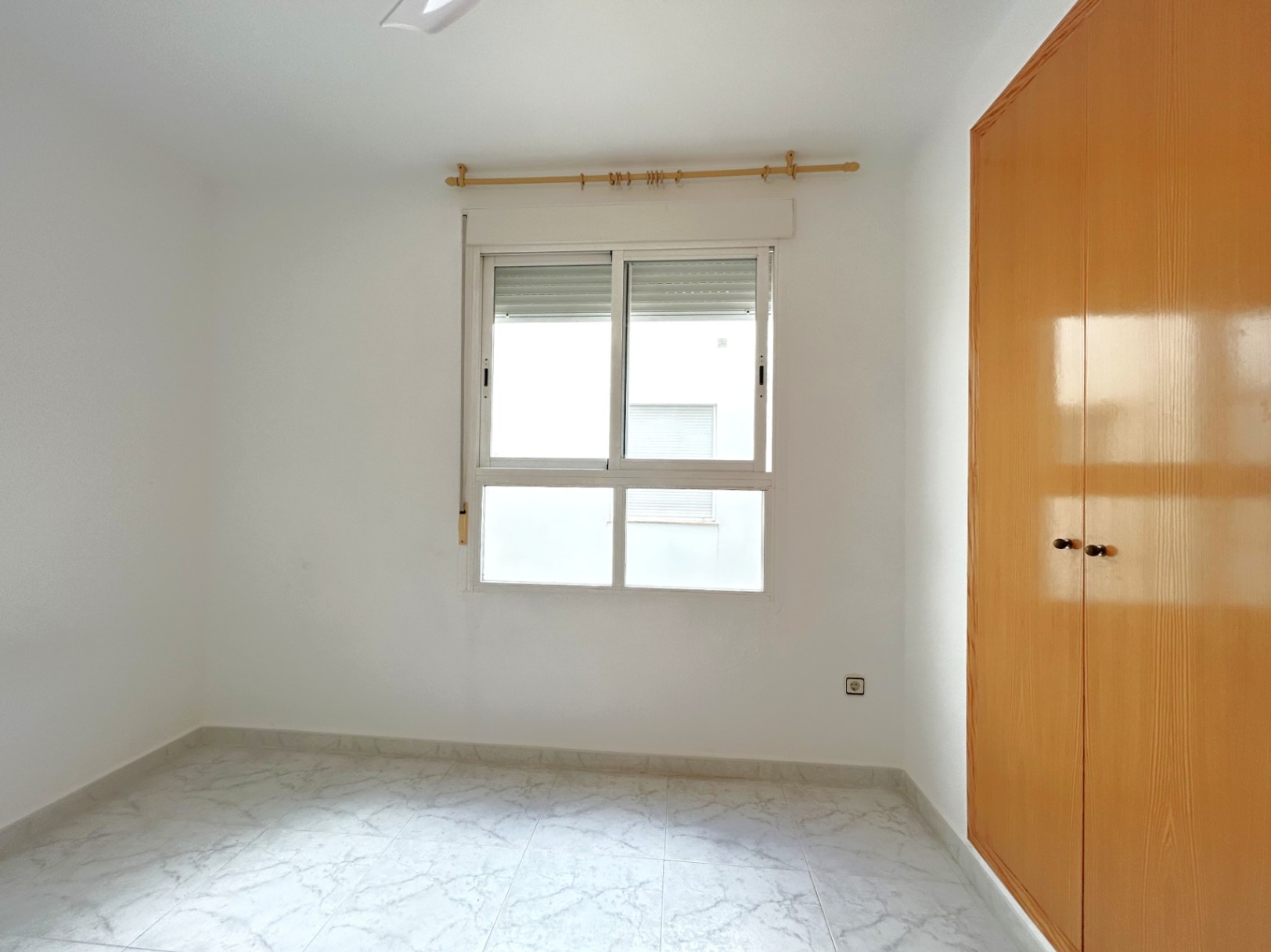 Chambre lumineuse avec sol en grès effet marbre, armoire encastrée en bois et fenêtre en aluminium blanc avec volet roulant.