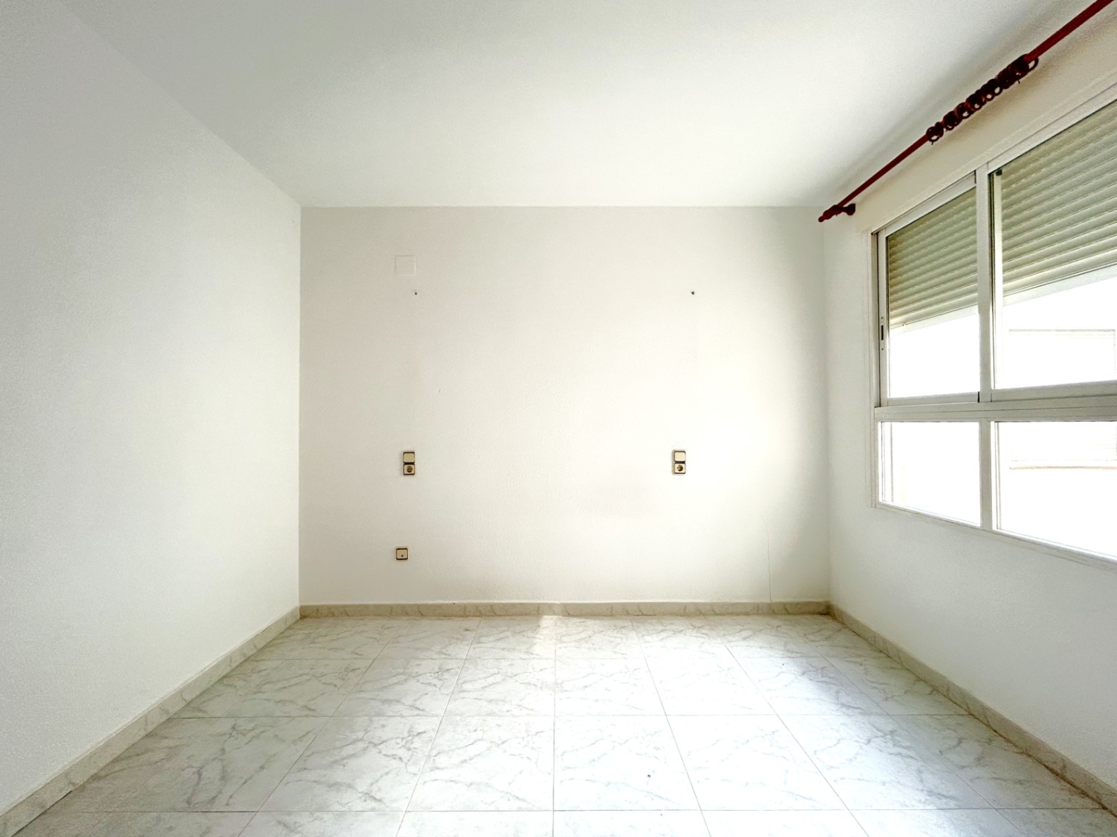 Chambre vide avec sol en grès cérame effet marbre, murs blancs et fenêtre en aluminium avec volet roulant. Prises électriques disposées pour l'emplacement du lit.