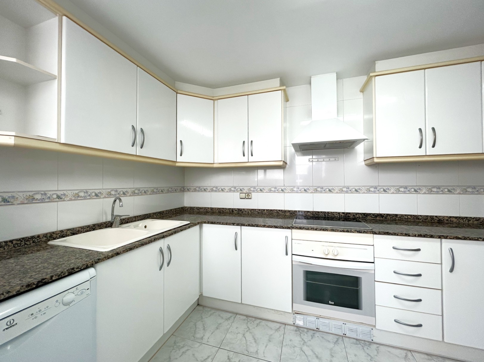 Cuisine indépendante avec placards blancs, plan de travail en granit, plaque vitrocéramique et four encastré. Les murs sont carrelés avec une frise décorative.