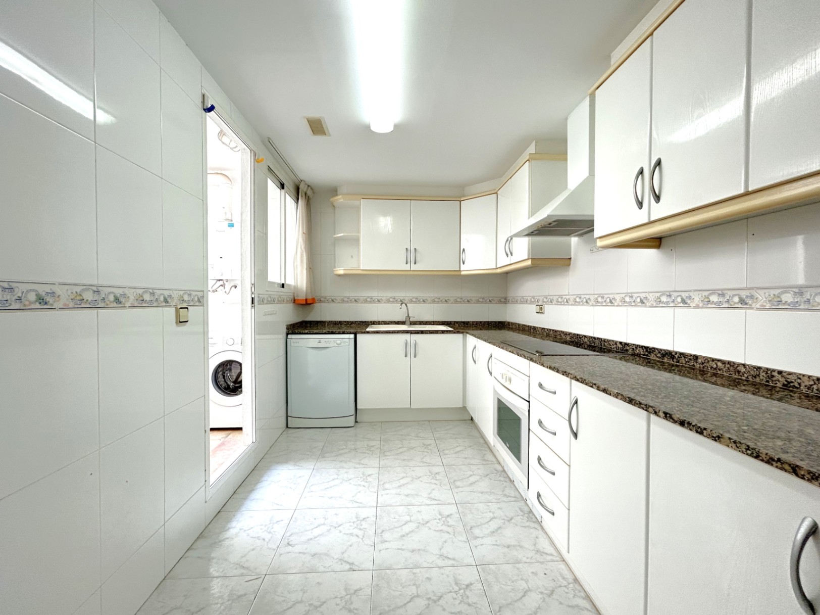 Cuisine indépendante en L avec meubles blancs, plans de travail en granit sombre et accès à une buanderie extérieure attenante.
