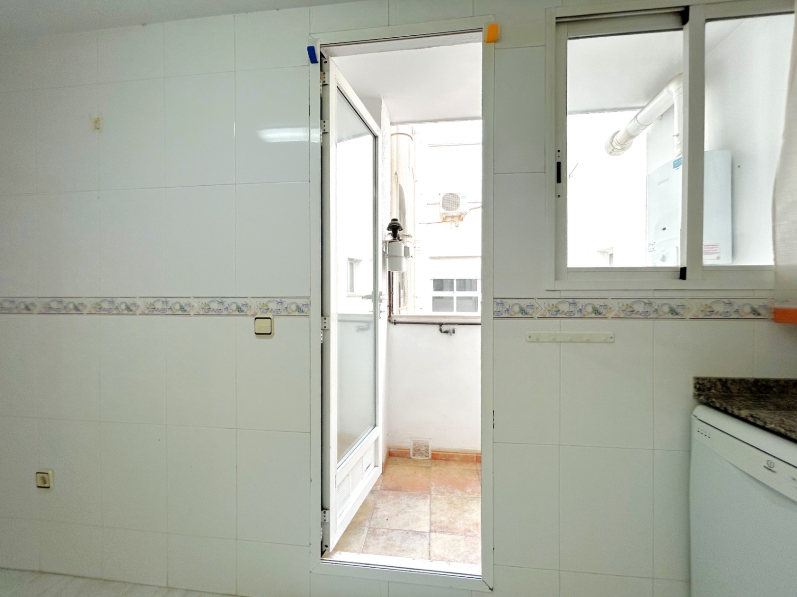 Galerie extérieure accessible depuis la cuisine, avec murs carrelés en blanc, plan de travail en granit et espace buanderie avec chauffe-eau.