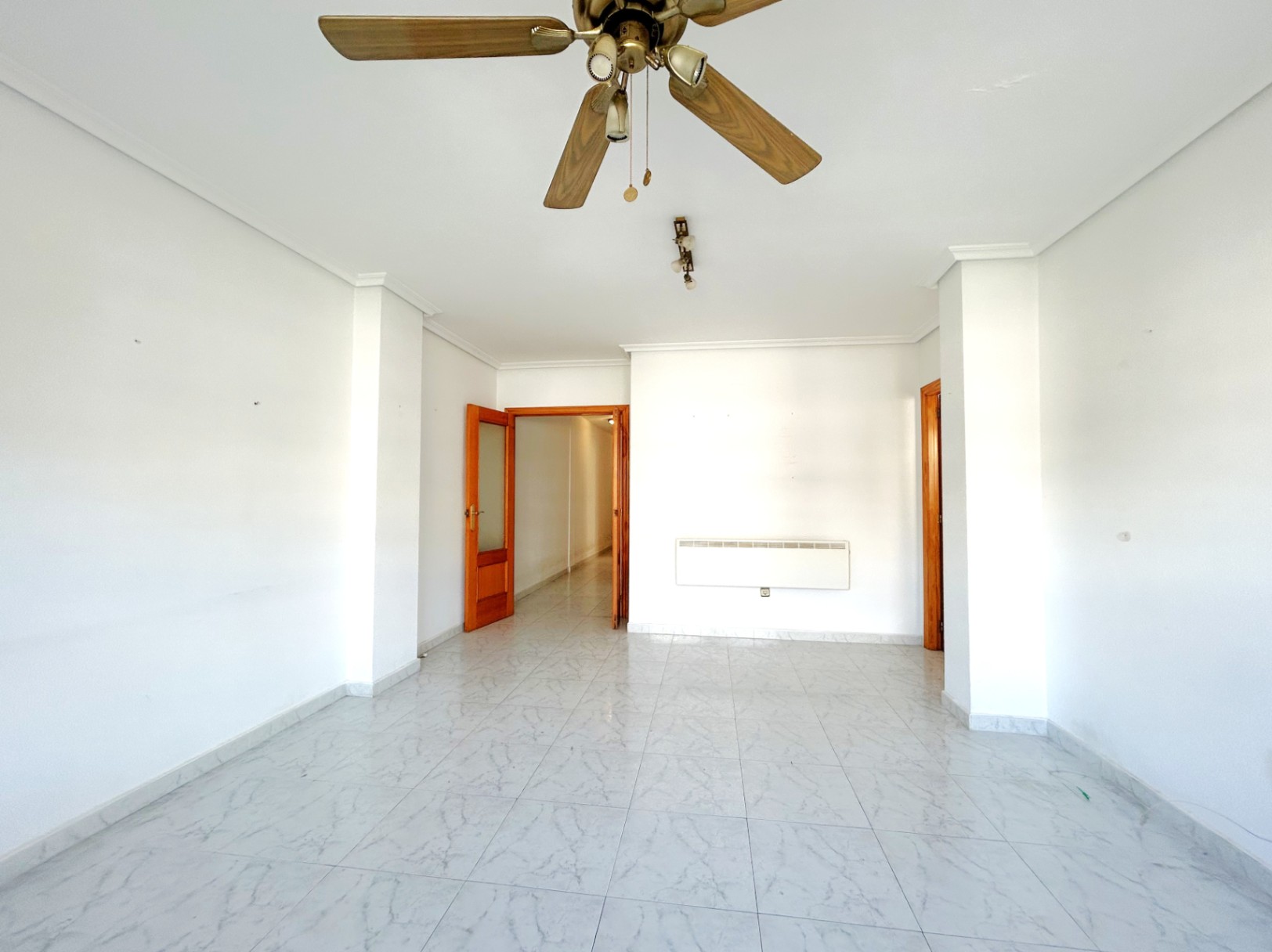 Grand salon lumineux avec sol en grès effet marbre, murs blancs et ventilateur de plafond en bois. Équipé d'un radiateur mural et de menuiseries intérieures en bois.