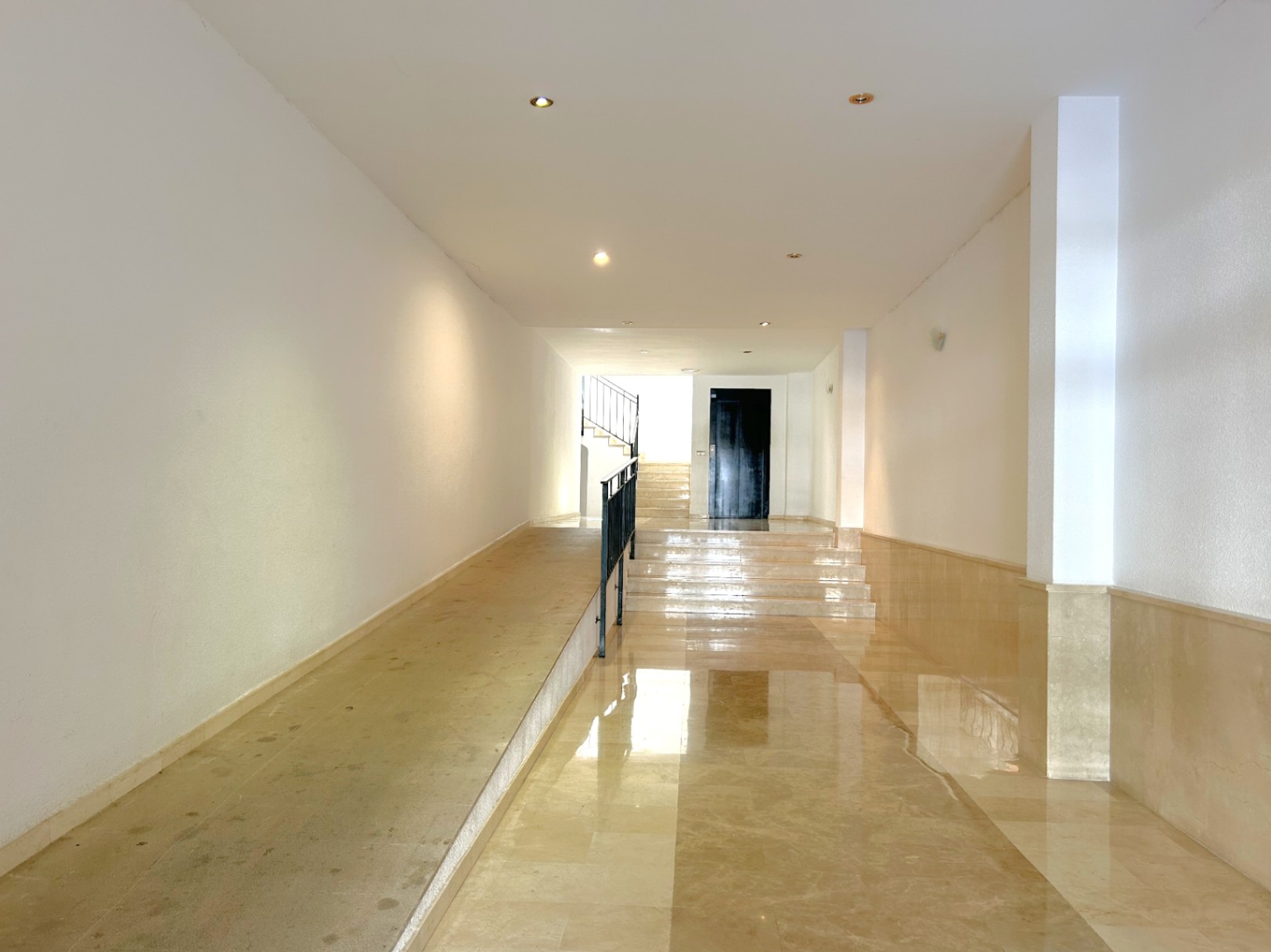 Hall d'entrée avec sol en marbre poli, rampe d'accès avec garde-corps métallique, éclairage encastré et murs blancs offrant une grande clarté.