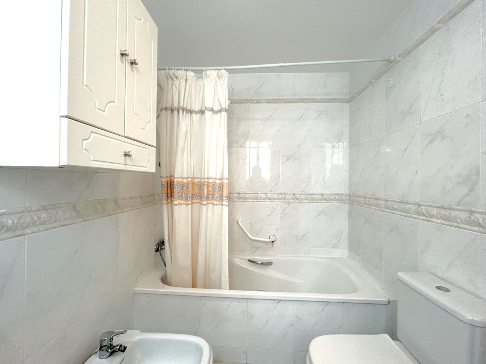 Salle de bain avec baignoire intégrée, carrelage effet marbre et frise décorative. Équipée d'un bidet, de toilettes et d'un meuble de rangement mural blanc.