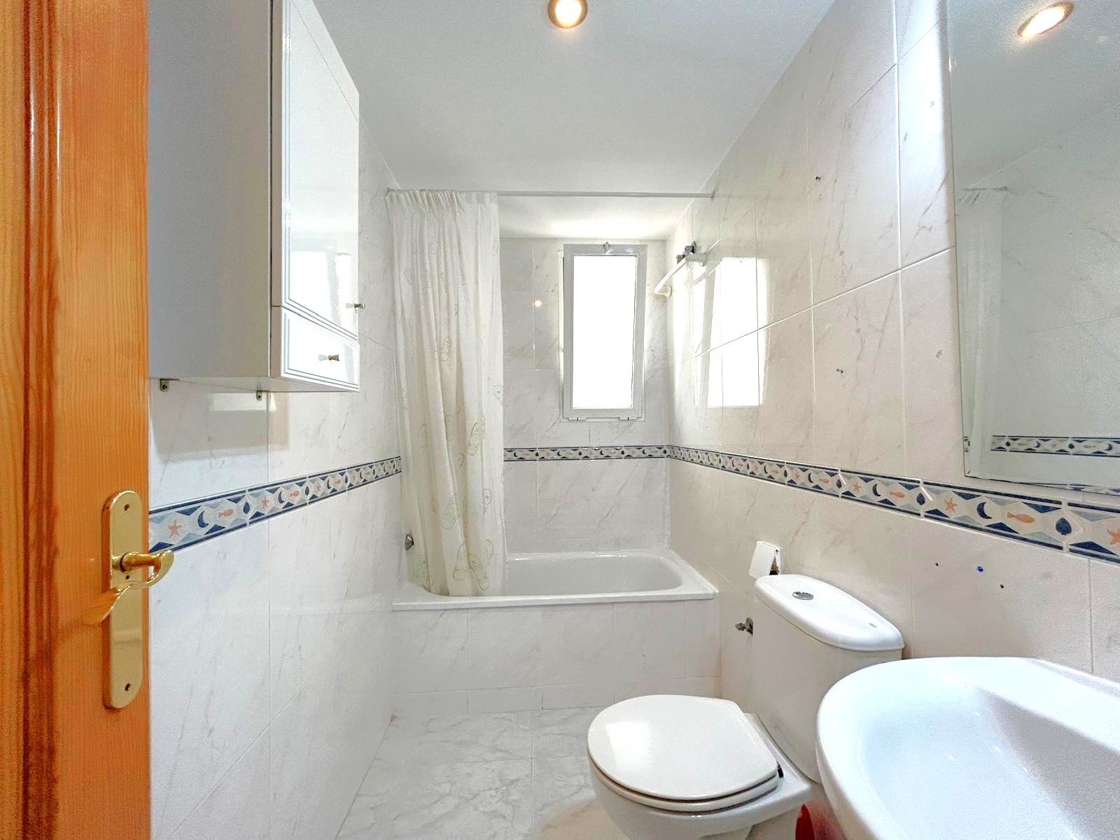 Salle de bain complète avec baignoire, carrelage effet marbre et frise décorative. Elle dispose d'une fenêtre pour la lumière naturelle et de rangements blancs.