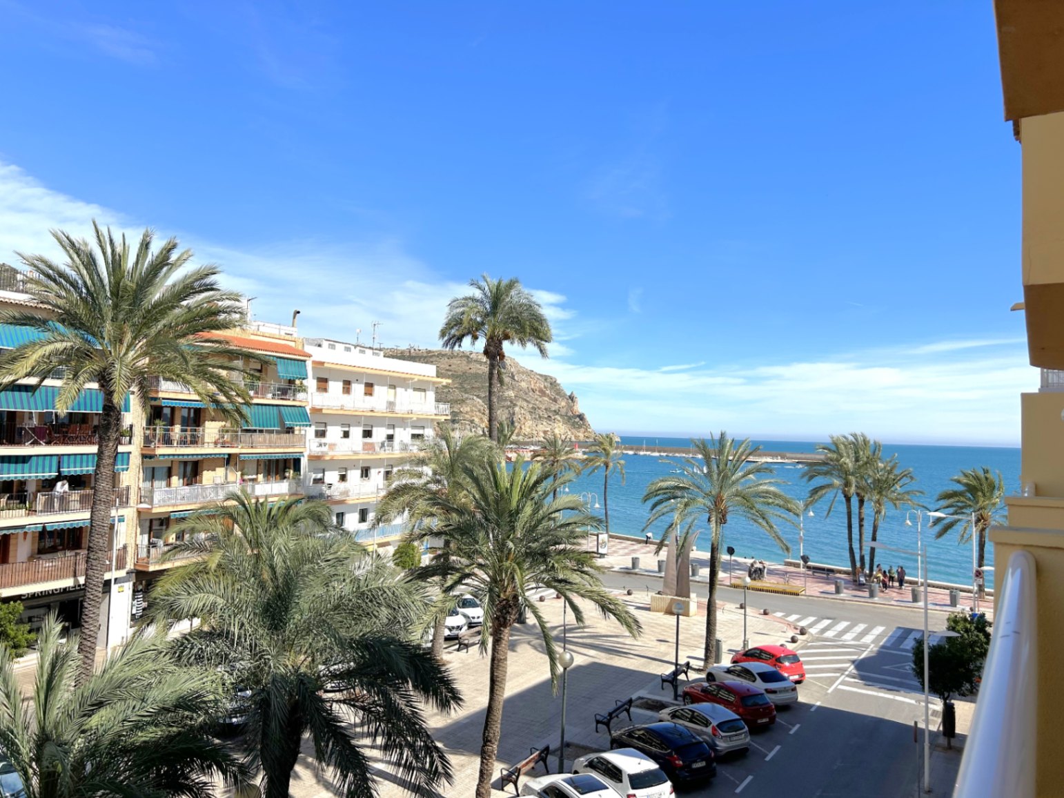 Vue depuis le balcon sur la promenade du port de Jávea, incluant des palmiers, la mer Méditerranée et les falaises environnantes par temps clair.