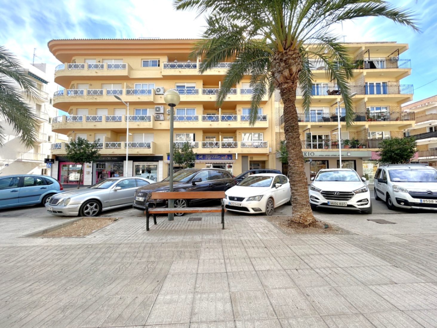 Vue extérieure du bâtiment au Port de Jávea, présentant une façade ocre et des balcons aux garde-corps blancs face à une place avec des palmiers.