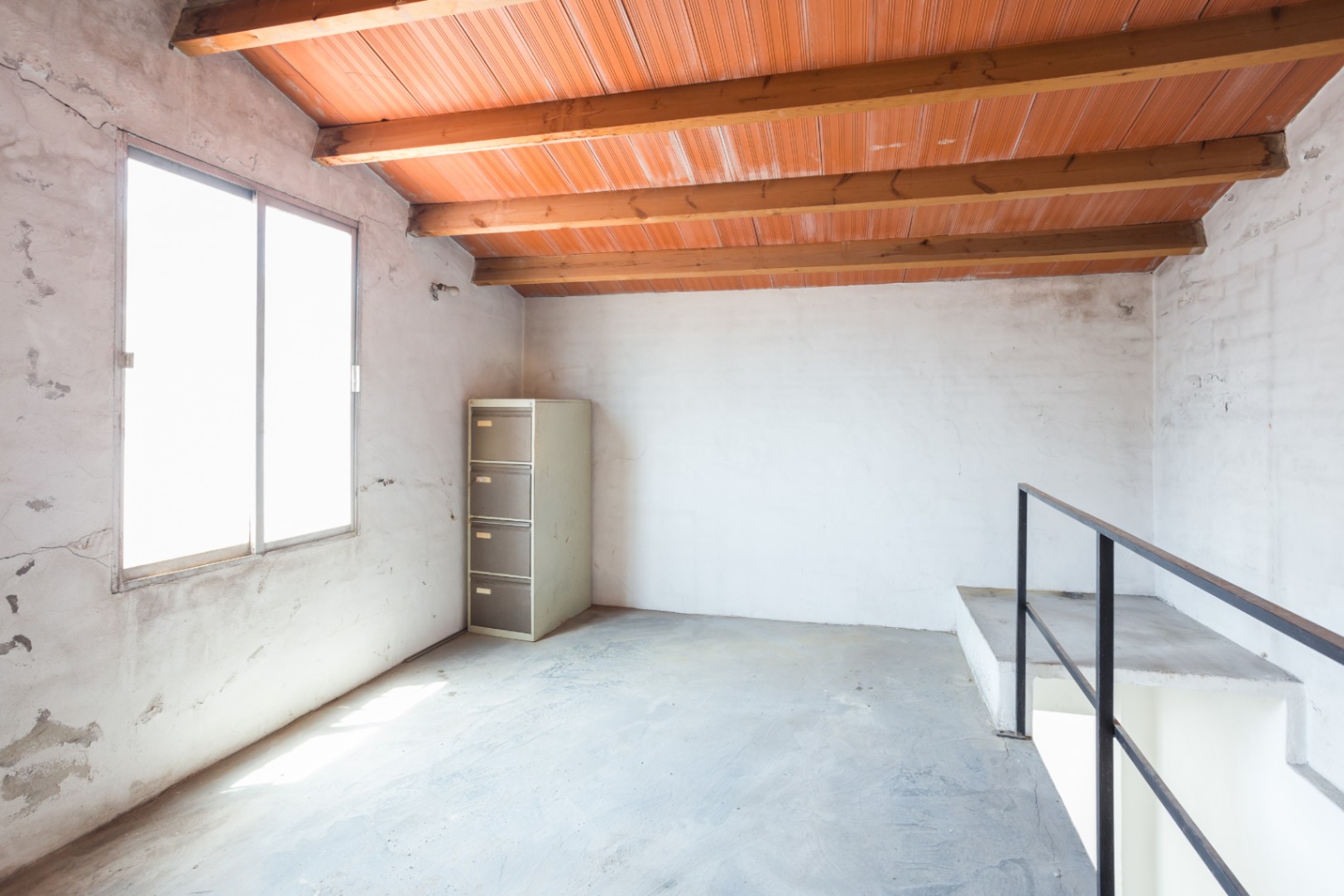 Étage supérieur avec poutres en bois apparentes, plafond en briques, sol en béton et une grande fenêtre offrant une lumière naturelle abondante.