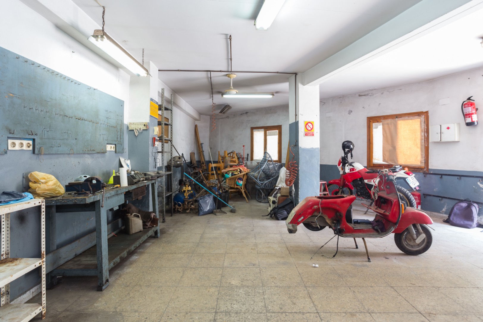 Grand atelier ou garage avec établi, étagères métalliques et sol carrelé, éclairé par des néons et bénéficiant d'une ventilation naturelle.