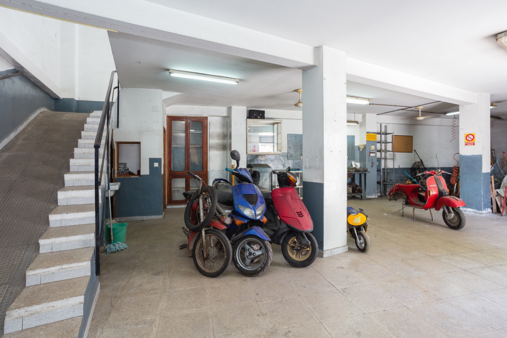 Grand espace ouvert de garage ou atelier avec piliers structurels, éclairage fluorescent et accès par escalier et rampe latérale.