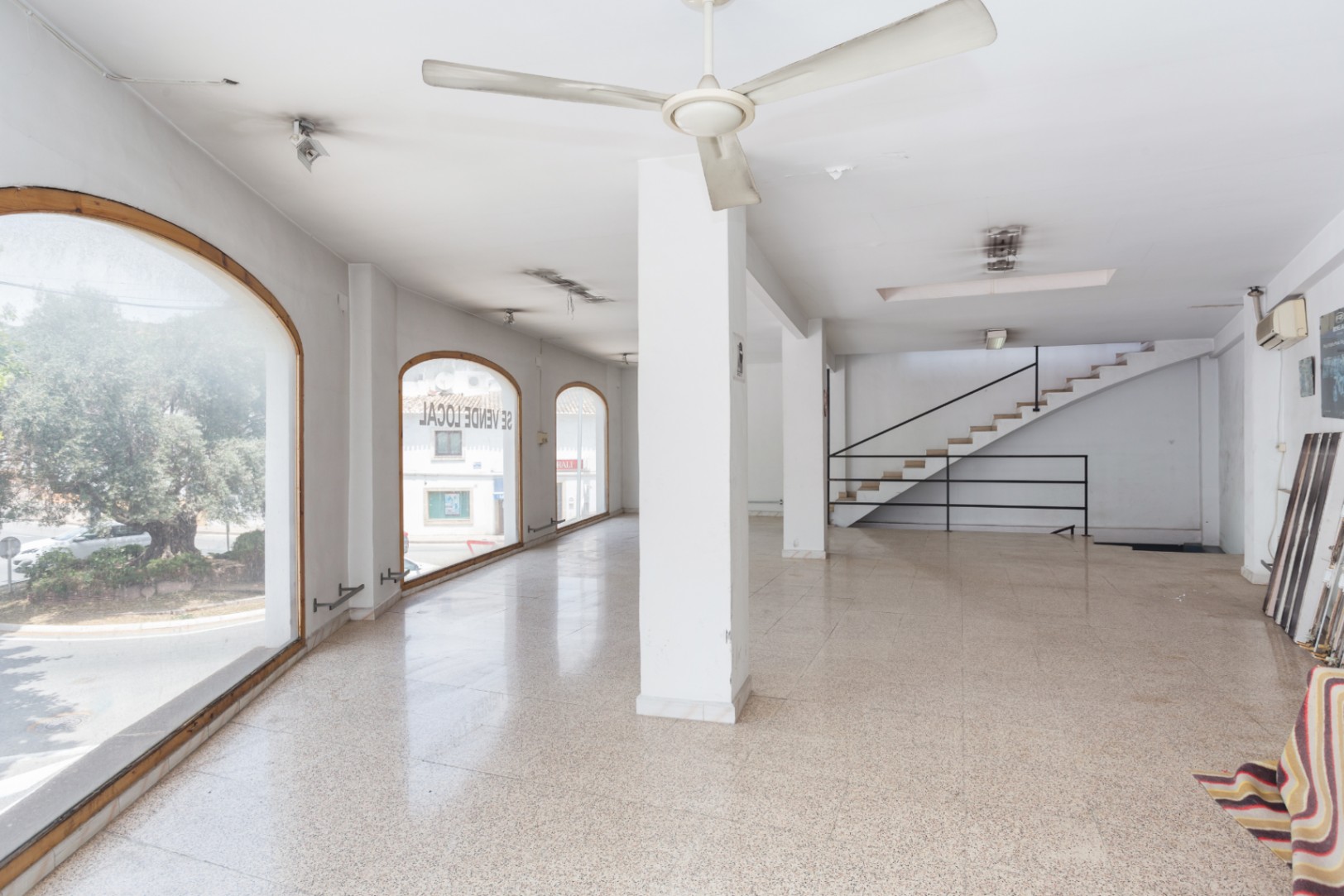 Rez-de-chaussée spacieux avec de grandes fenêtres cintrées, sol en terrazzo et un escalier avec rampe en métal noir menant à l'étage.