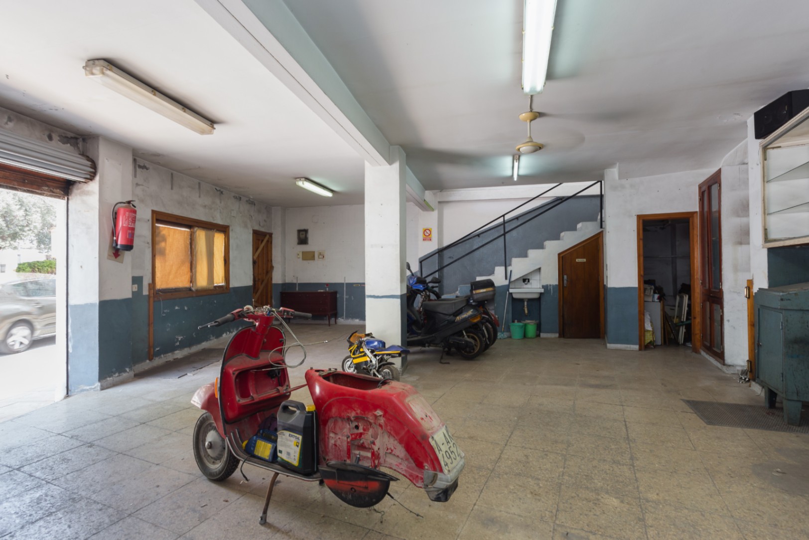 Rez-de-chaussée spacieux avec sol en céramique, accès par rideau métallique et escalier vers l'étage. Espace polyvalent avec éclairage fluorescent.