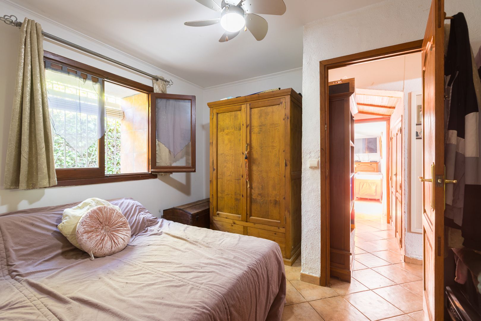 Chambre chaleureuse avec armoire en bois massif, sol en grès et ventilateur de plafond. La fenêtre à cadre en bois laisse entrer la lumière naturelle sur le lit.