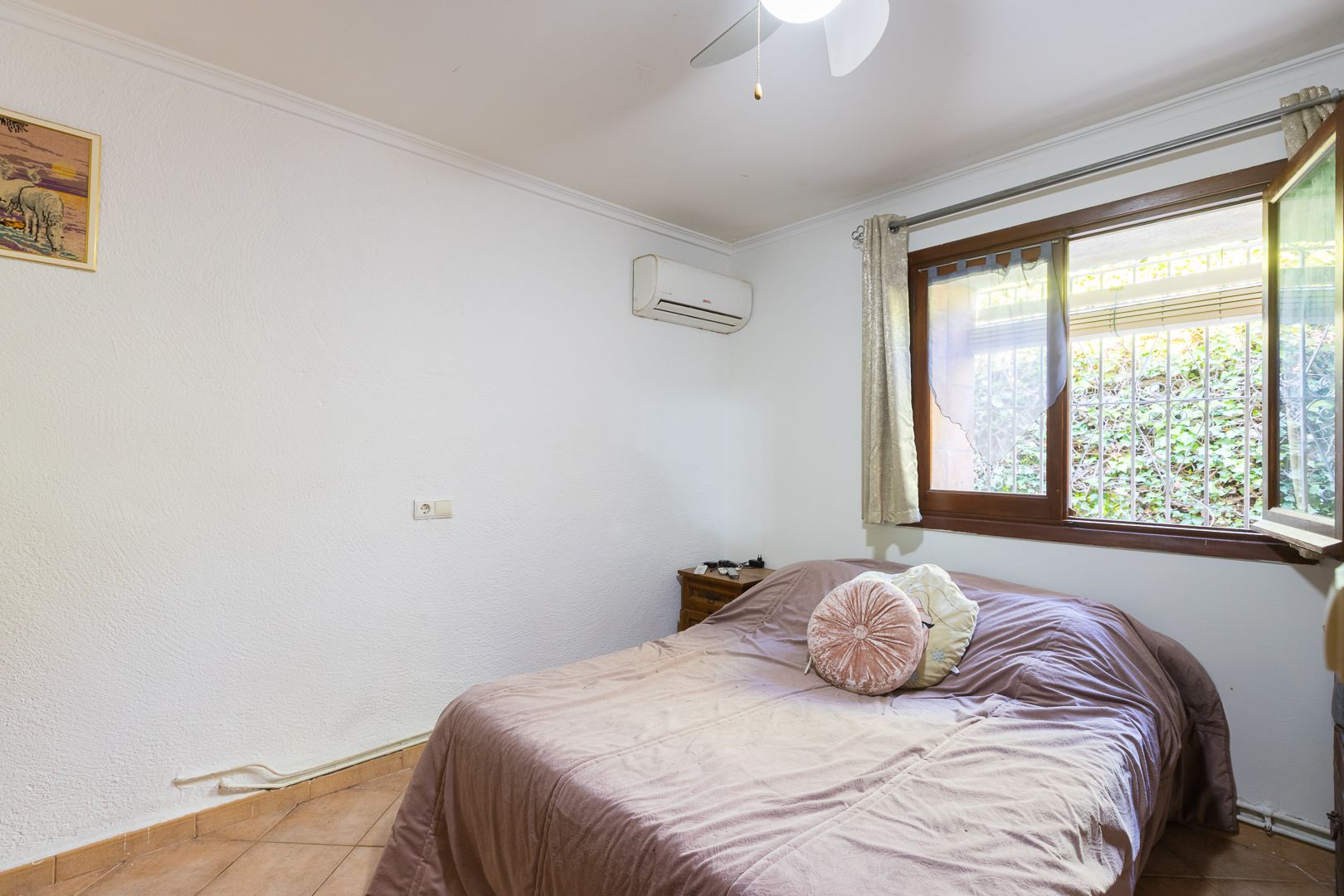 Chambre lumineuse équipée d'une climatisation split, d'un ventilateur de plafond et d'une fenêtre en bois avec vue sur la végétation.