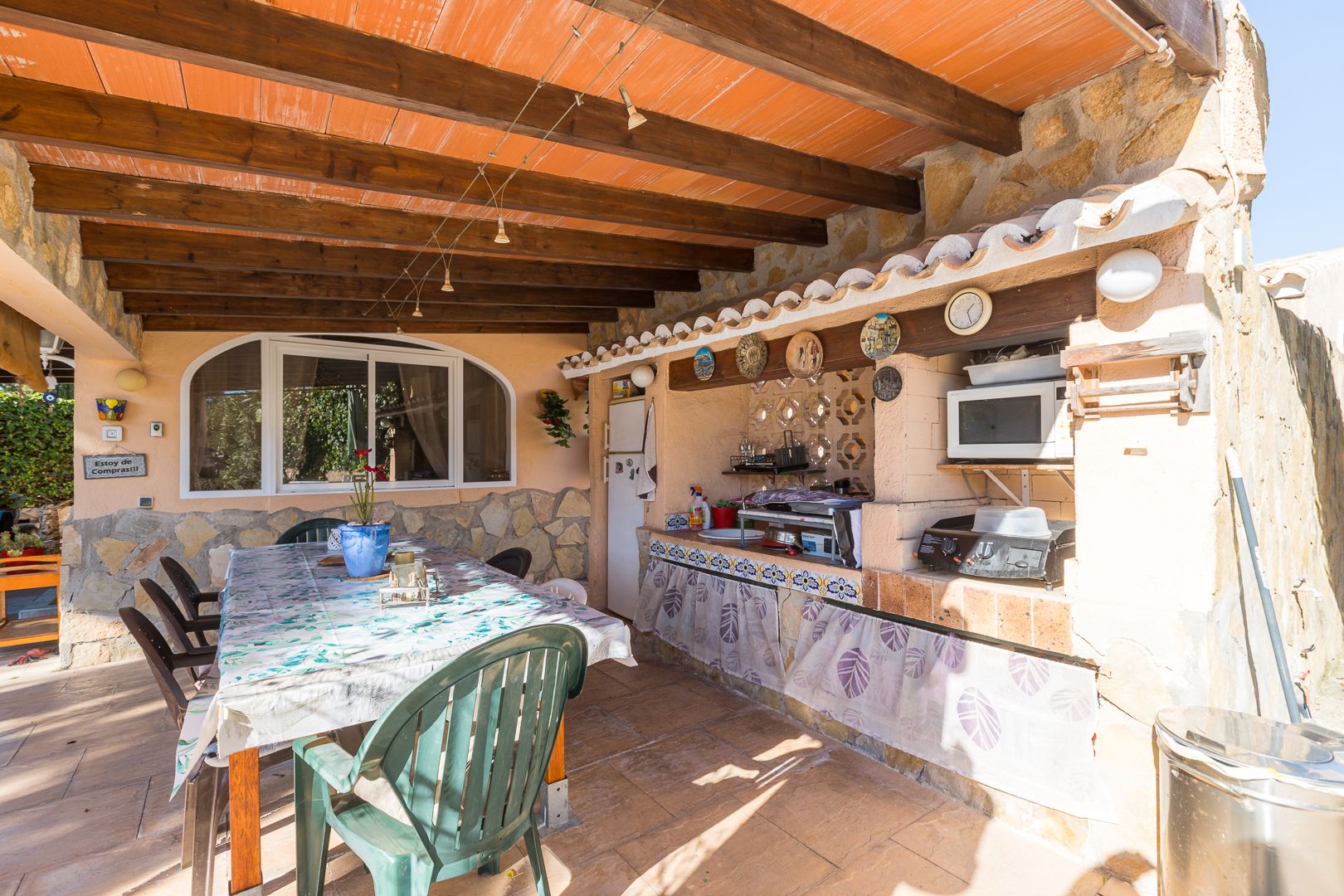 Cuisine d'été et espace repas sous une terrasse couverte avec poutres en bois, murs en pierre naturelle et coin barbecue aménagé.