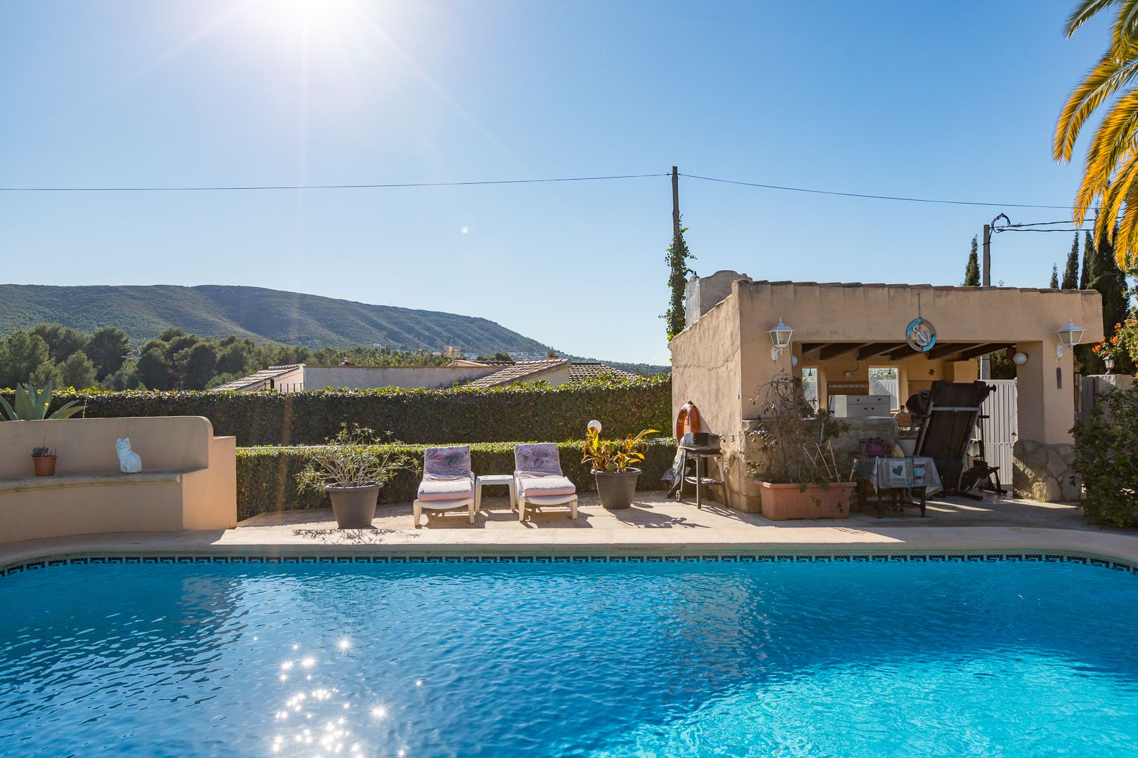 Piscine avec bordure en mosaïque, terrasse ensoleillée avec transats et cuisine d'été couverte, offrant une vue dégagée sur les montagnes environnantes.