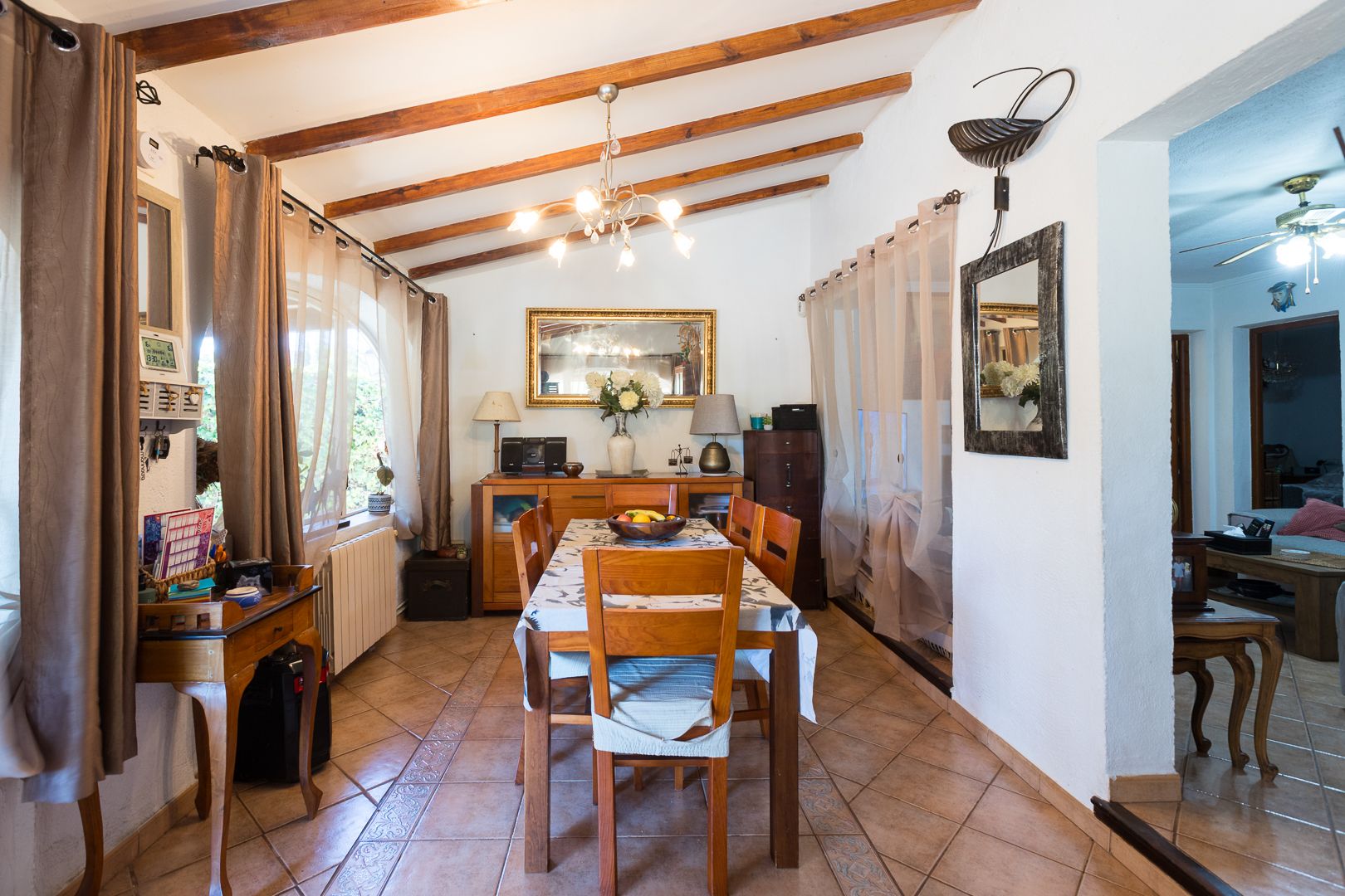Salle à manger lumineuse avec plafond incliné à poutres apparentes, sol en grès rustique et mobilier en bois, offrant une atmosphère méditerranéenne chaleureuse.