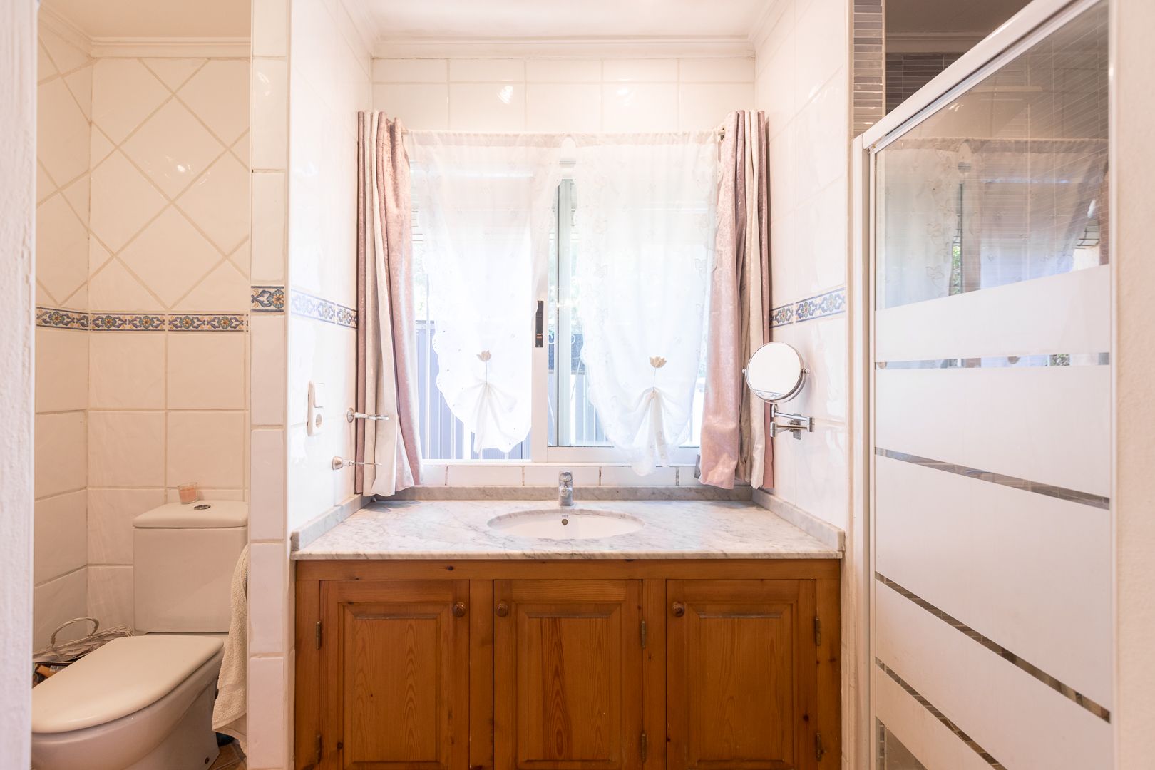 Salle de bain avec meuble sous-vasque en bois, plan de travail en marbre et fenêtre. Comprend une paroi de douche sérigraphiée et des carreaux muraux décorés.