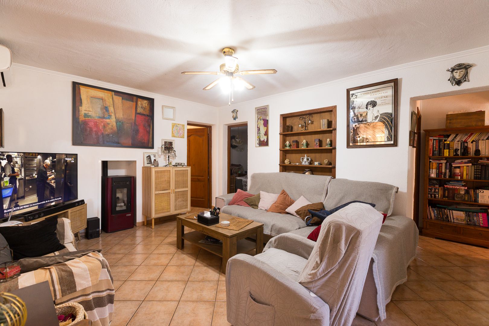 Salon avec sol en grès, poêle à granulés, ventilateur de plafond et étagères en bois intégrées, offrant un espace de vie confortable et fonctionnel.