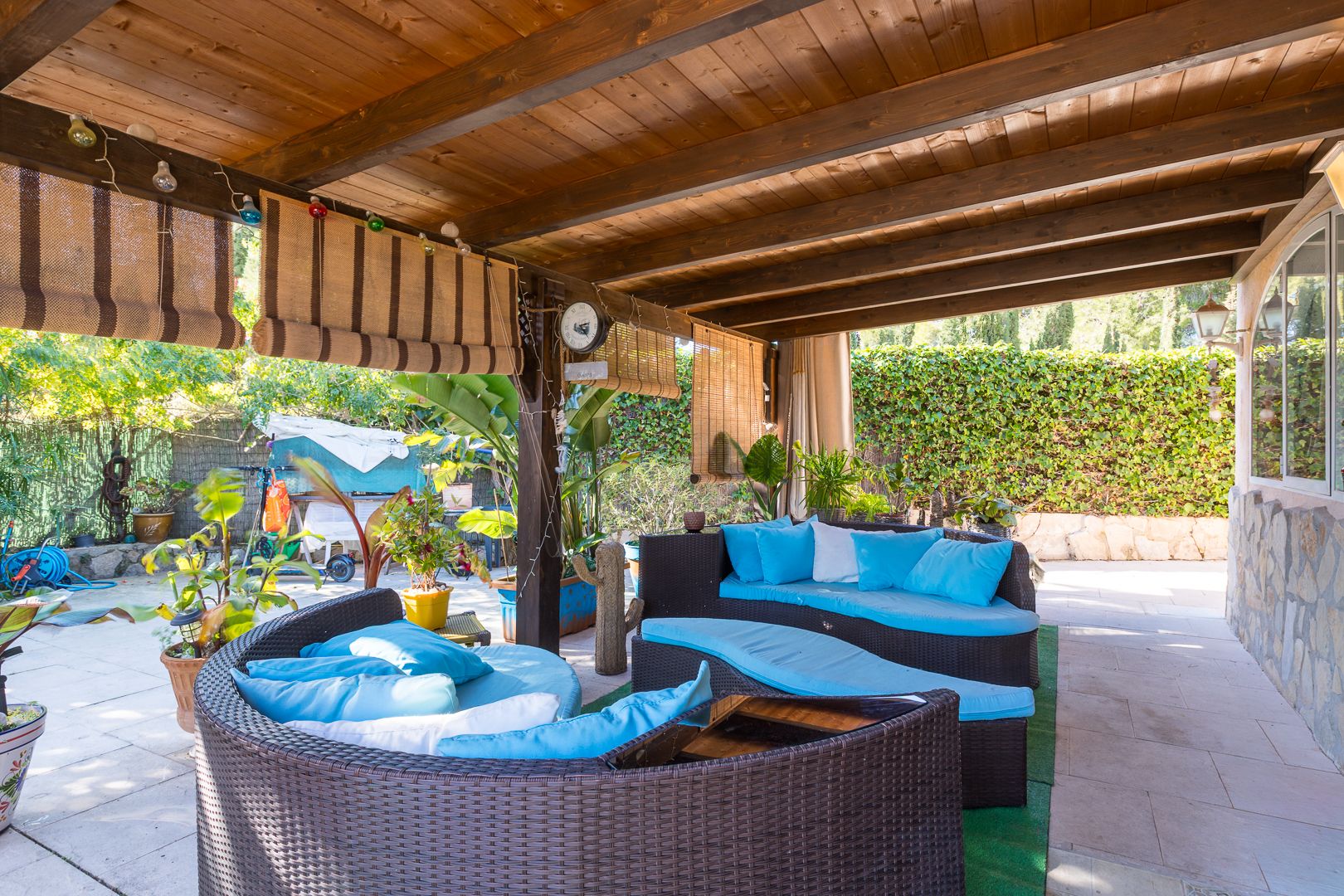 Terrasse couverte avec pergola en bois sombre, mobilier de salon en rotin arrondi aux coussins bleus, et stores en bambou offrant ombre et intimité.