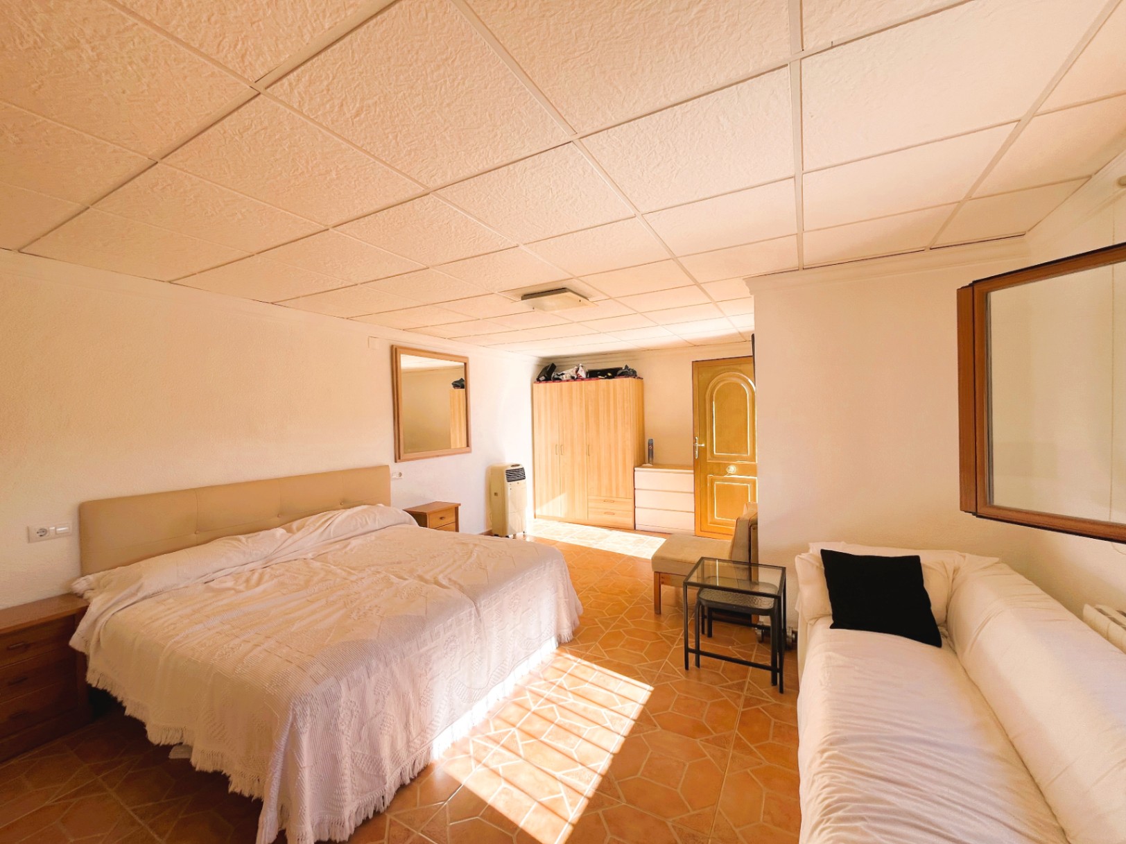 Chambre avec sol en terre cuite, faux plafond modulaire et coin salon. Le mobilier en bois est mis en valeur par une lumière naturelle abondante.
