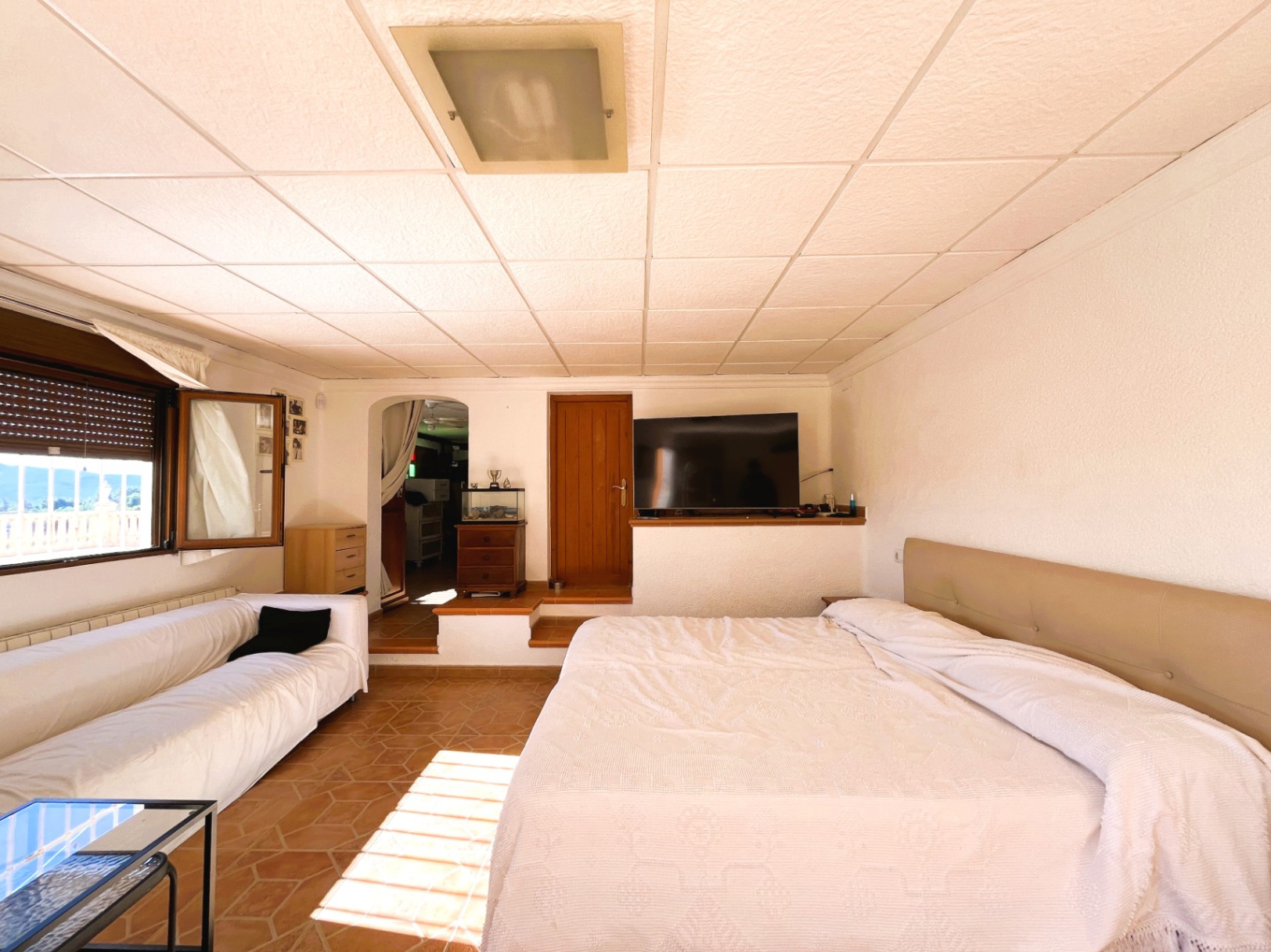 Chambre en rez-de-jardin avec sol en terre cuite, espace salon intégré et fenêtre offrant une vue dégagée sur les montagnes environnantes.