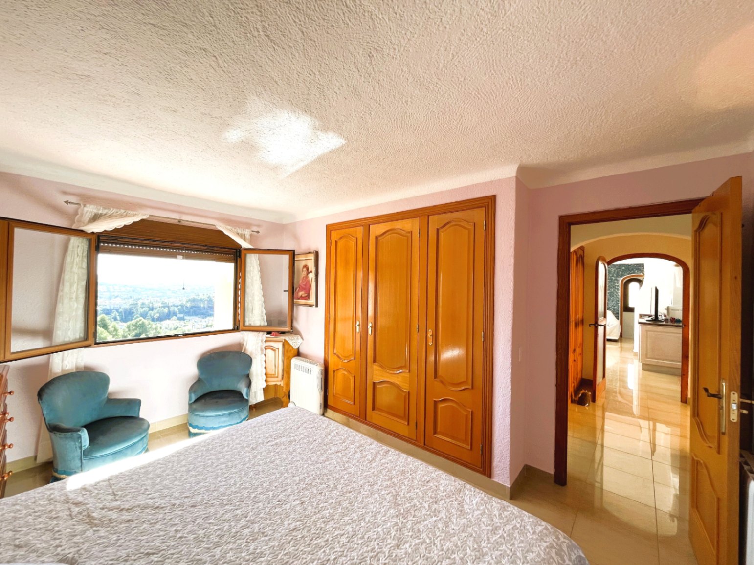 Chambre lumineuse avec placards intégrés en bois, fauteuils en velours et fenêtre offrant une vue panoramique dégagée sur les environs.