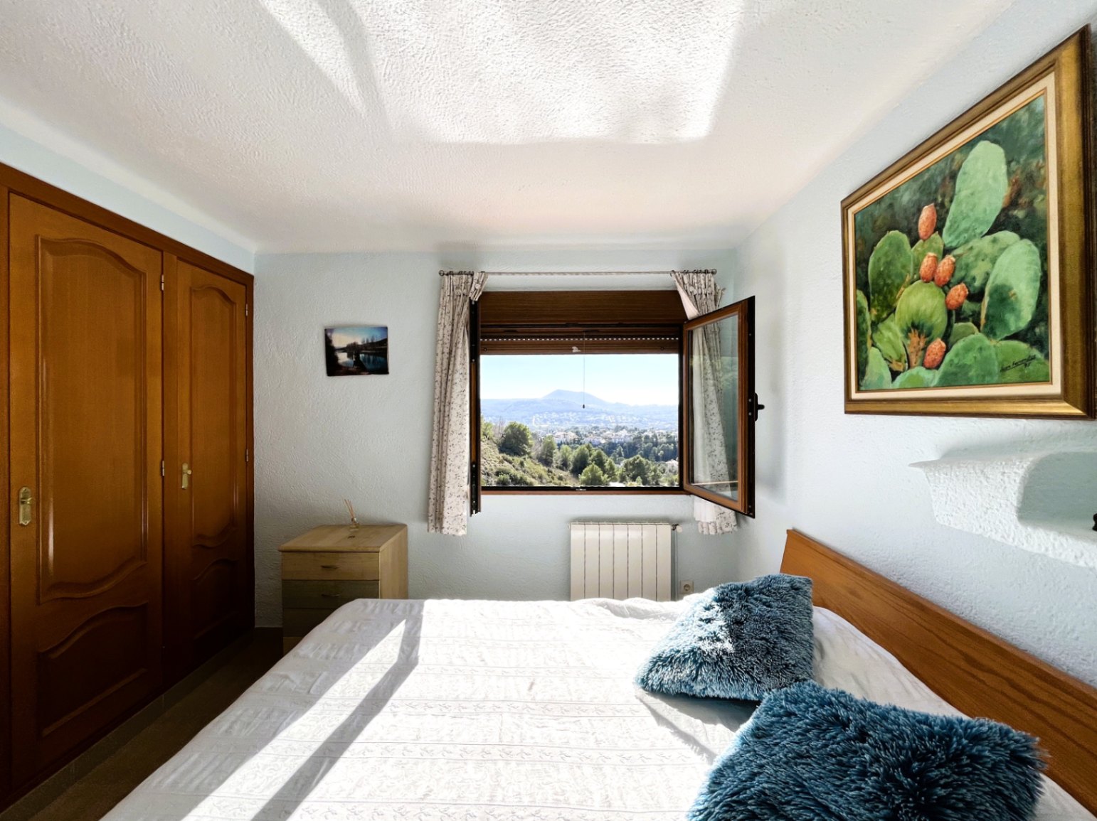 Chambre lumineuse avec placards intégrés en bois, radiateur et fenêtre ouverte offrant une vue dégagée sur les montagnes et la nature environnante.