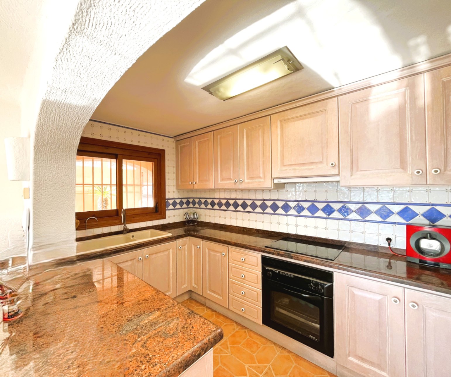 Cuisine équipée avec des meubles en bois clair, un plan de travail en granit et une crédence en carrelage aux motifs bleus. Elle dispose d'un four intégré et d'une plaque vitrocéramique.
