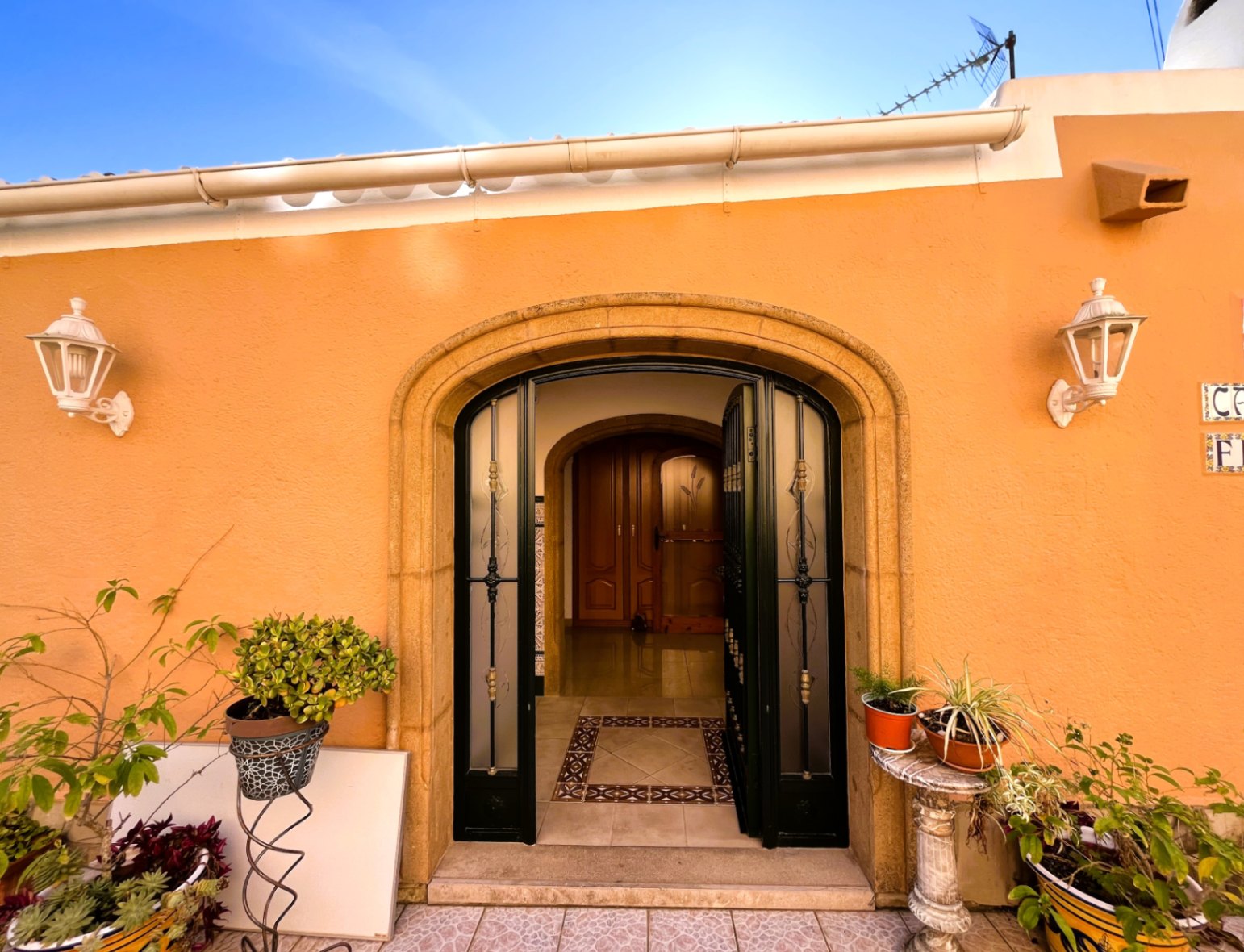 Entrée principale avec arche en pierre naturelle, porte en fer forgé et verre dépoli, sur une façade ocre ornée de lanternes blanches et de plantes en pot.