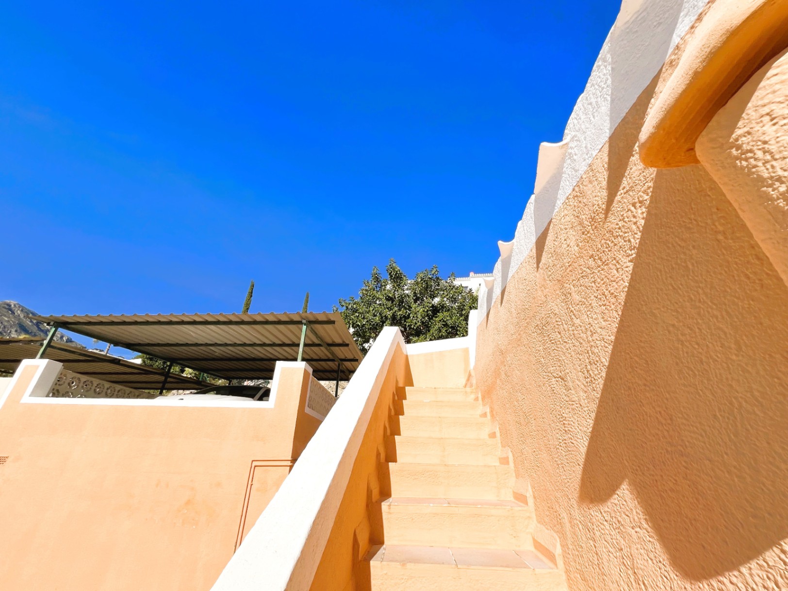 Escalier extérieur carrelé bordé de murs aux tons terre et blanc, situé à côté de l'espace de stationnement couvert par une pergola métallique.