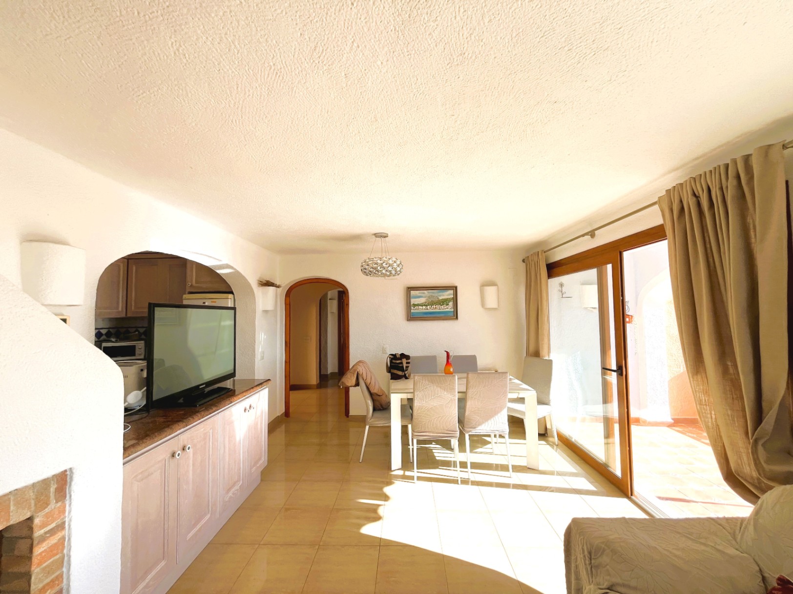 Salle à manger lumineuse avec sol en grès, grandes baies vitrées en bois ouvrant sur la terrasse et accès fluide vers la cuisine par une ouverture en arche.
