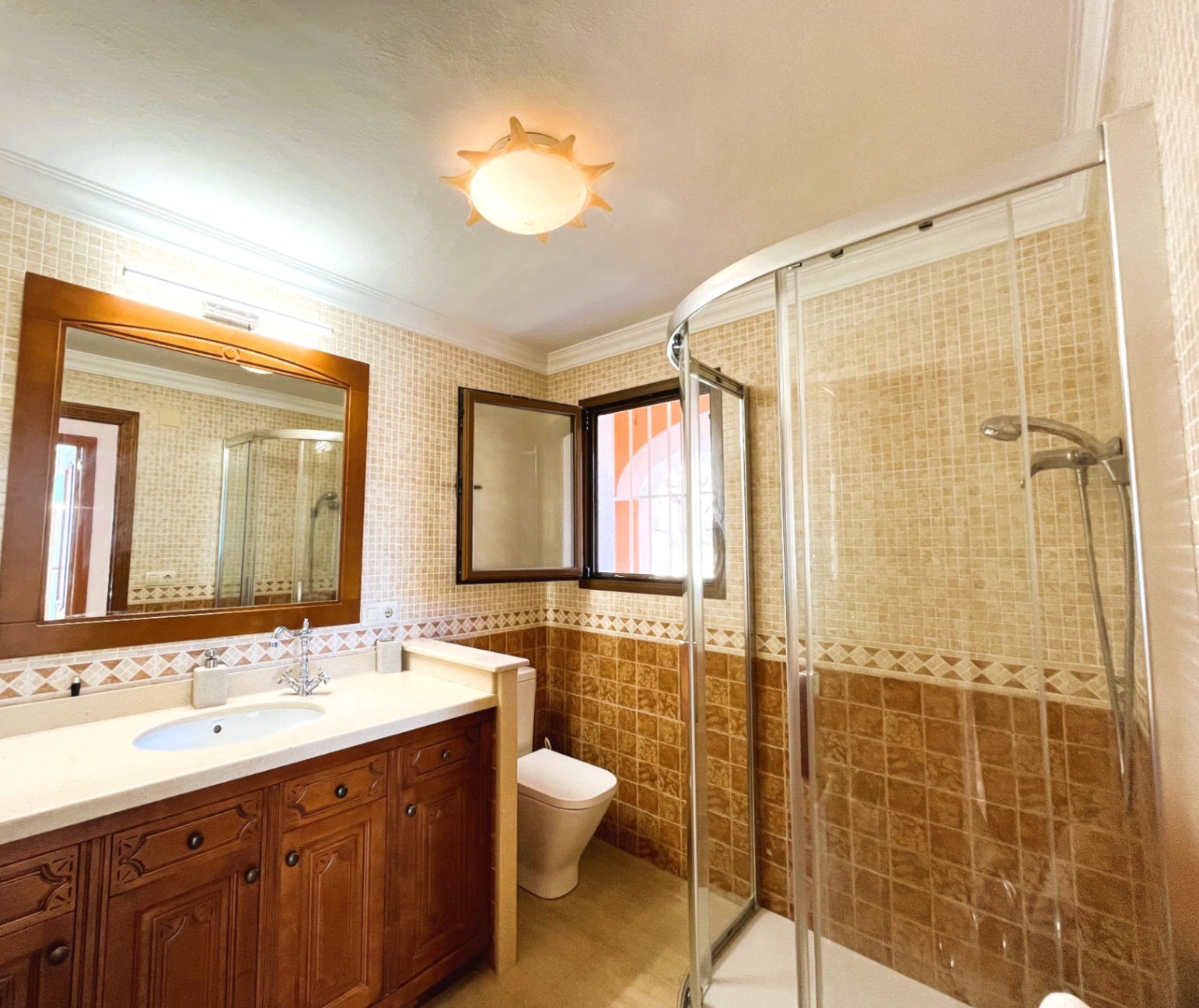 Salle de bain avec meuble en bois sculpté, douche d'angle à paroi vitrée courbe et carrelage mural aux tons beiges et ocres.