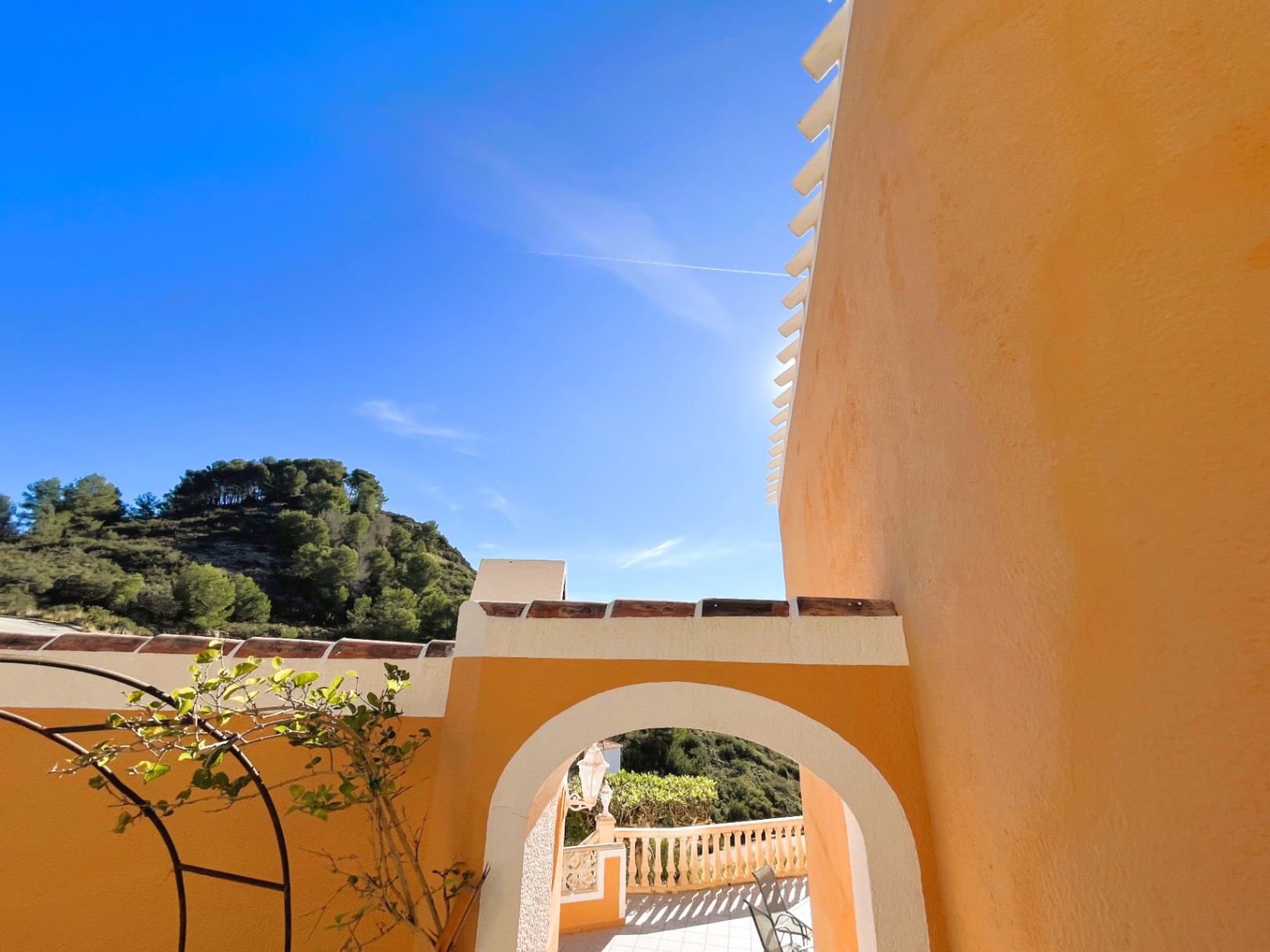 Vue extérieure montrant une arche d'entrée blanche sur une façade ocre, avec des tuiles en terre cuite et une vue sur les collines boisées.
