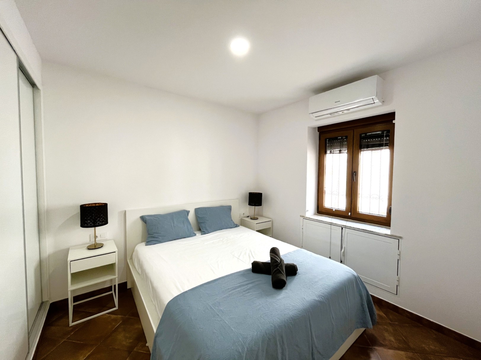 Chambre avec lit double, tables de chevet blanches et climatisation. Elle dispose d'un sol en carrelage rustique, d'un placard intégré et d'une fenêtre en bois.