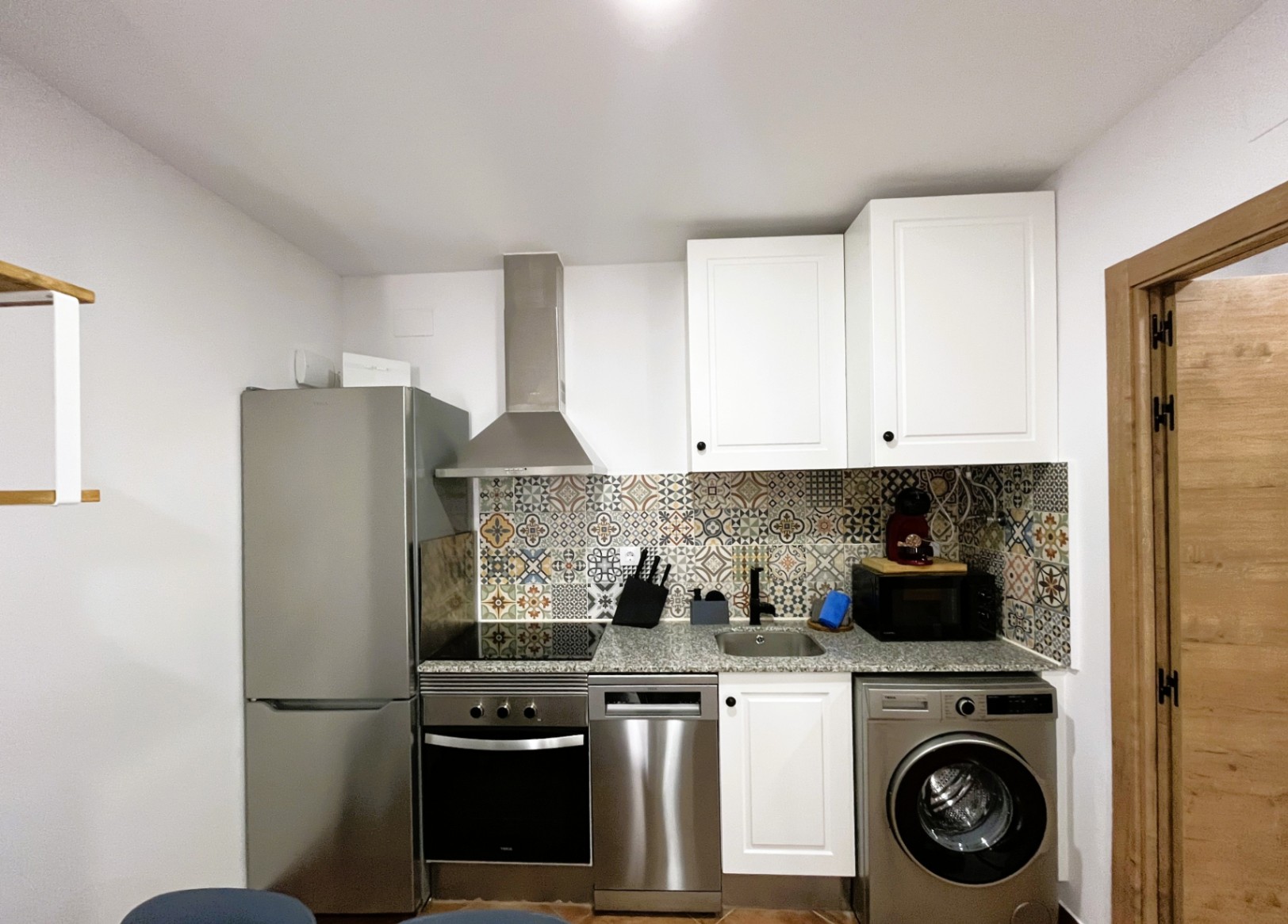 Cuisine compacte rénovée avec plan de travail en granit, crédence en carreaux de ciment et électroménagers en acier inoxydable, incluant lave-vaisselle et lave-linge.