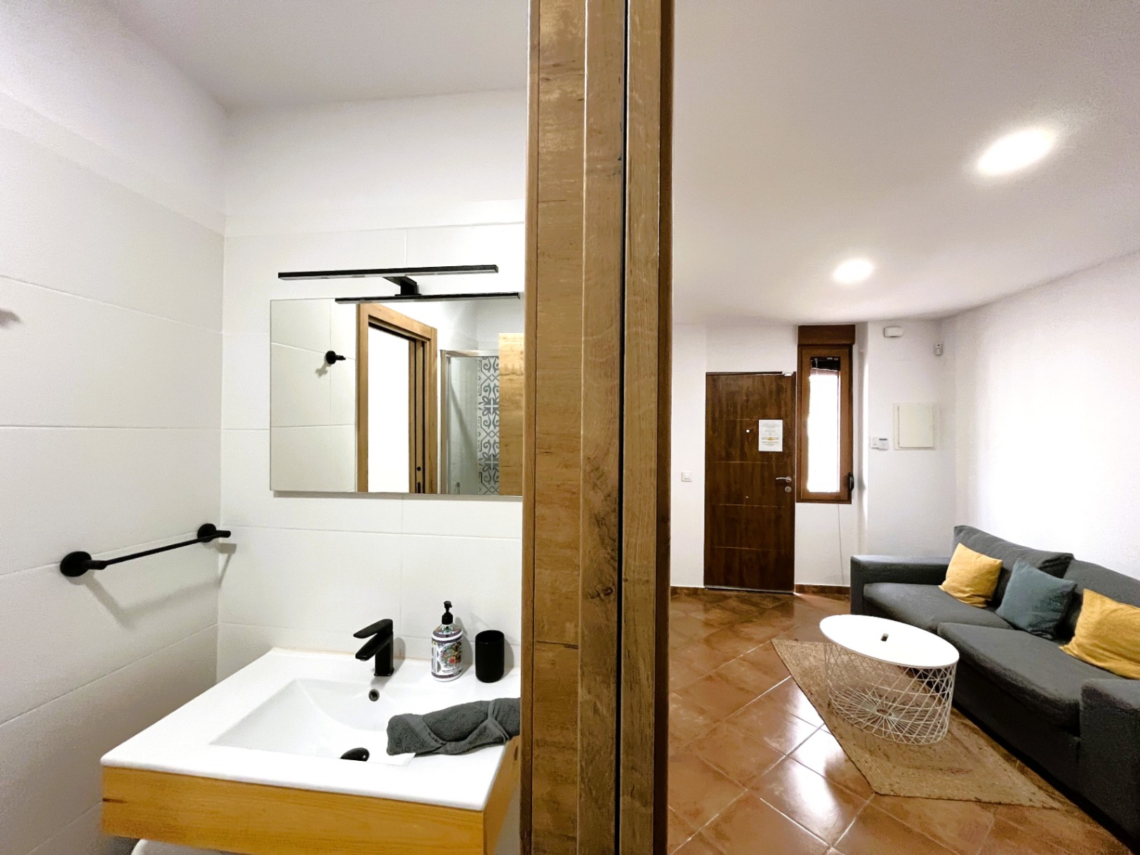 Espace de vie avec sol en terre cuite et canapé gris, à côté d'une salle de bain moderne avec vasque blanche, robinetterie noire et carrelage épuré.