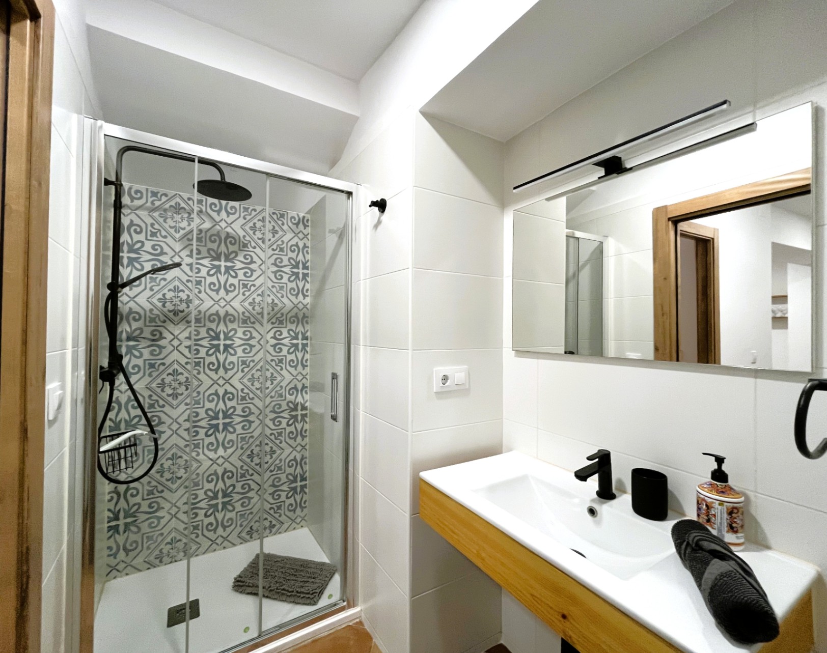 Salle de bain moderne avec douche à l'italienne, carrelage décoratif et robinetterie noire. Meuble vasque avec finitions en bois et miroir éclairé par un luminaire linéaire.