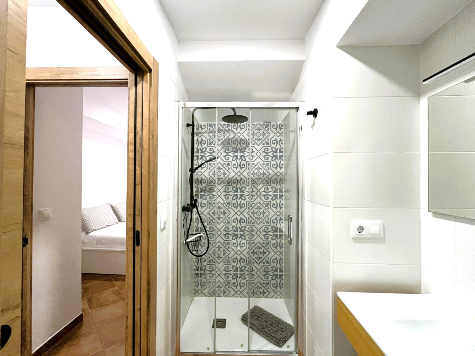 Salle de bain rénovée avec douche à l'italienne, carrelage hydraulique décoratif et robinetterie noire. Vue sur la chambre à travers le cadre de porte en bois.