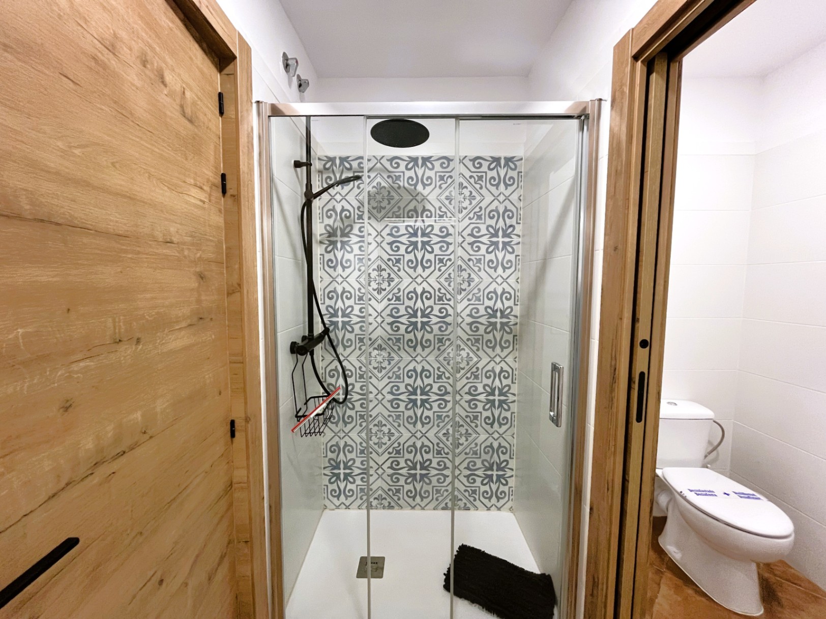 Salle de bain rénovée avec douche à l'italienne, parois en verre et carrelage décoratif à motifs. Robinetterie noire et portes en finition bois naturel.