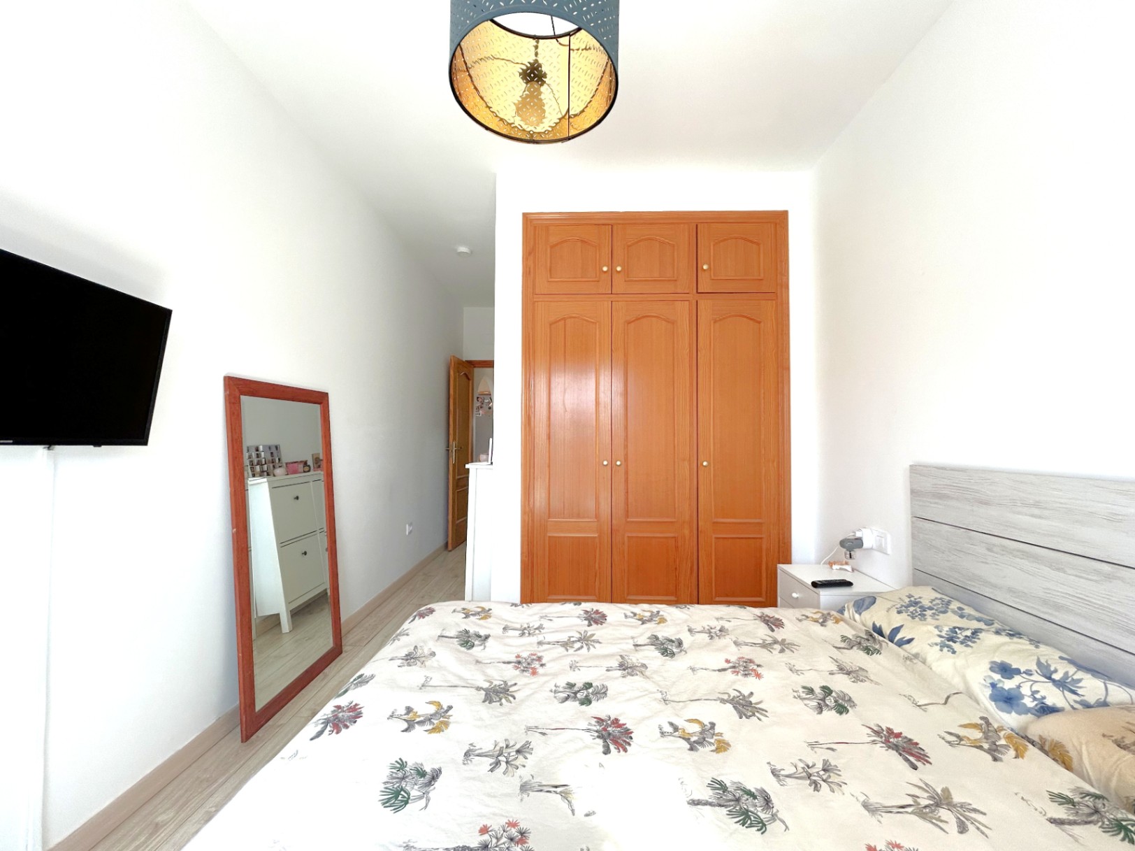 Chambre double avec armoire encastrée en bois, parquet clair et télévision murale. La pièce dispose d'un miroir sur pied et d'un luminaire décoratif au plafond.
