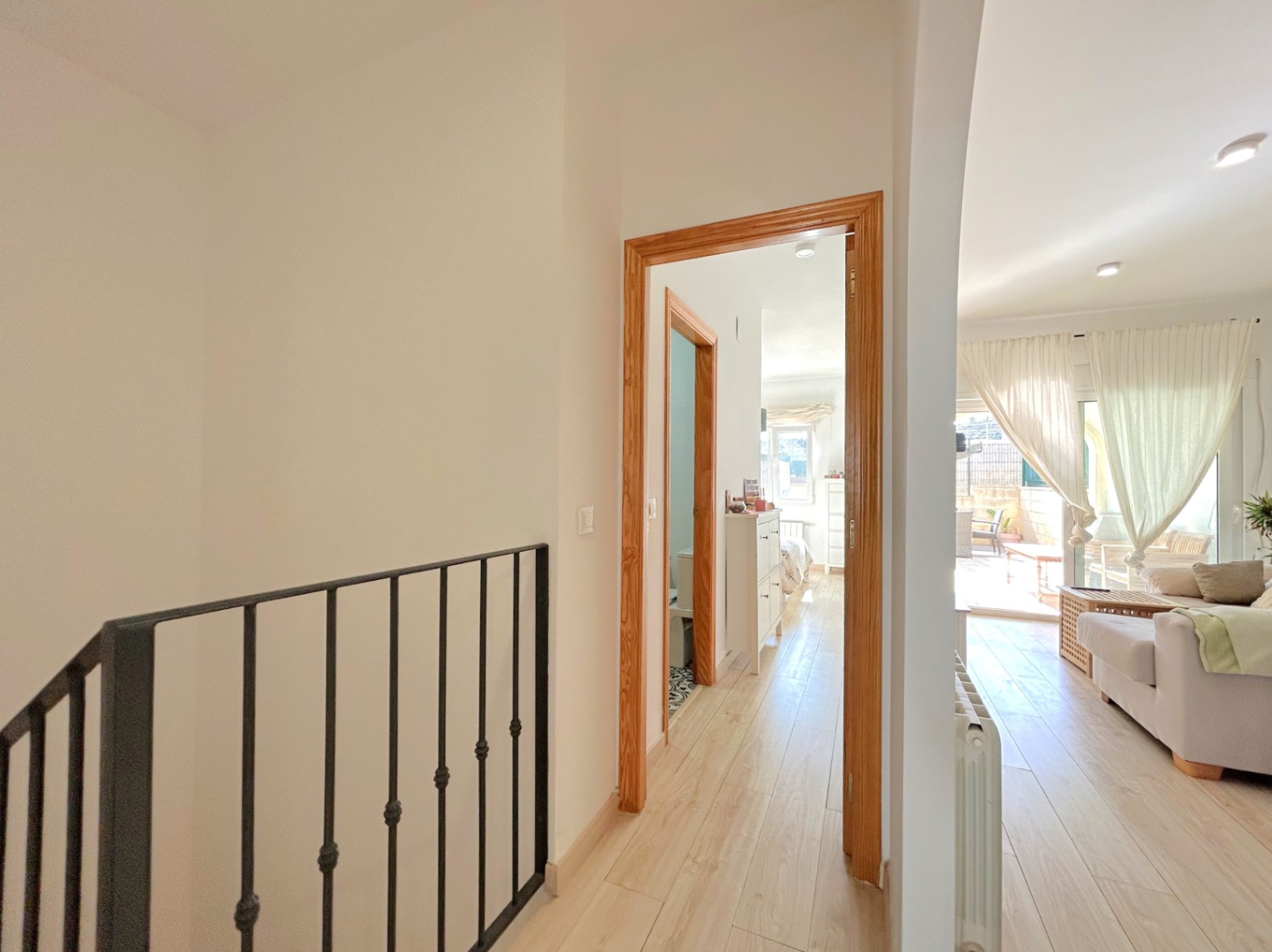 Couloir lumineux avec parquet clair et rampe en fer forgé, reliant le salon aux chambres et à la salle de bain, incluant un radiateur de chauffage central.