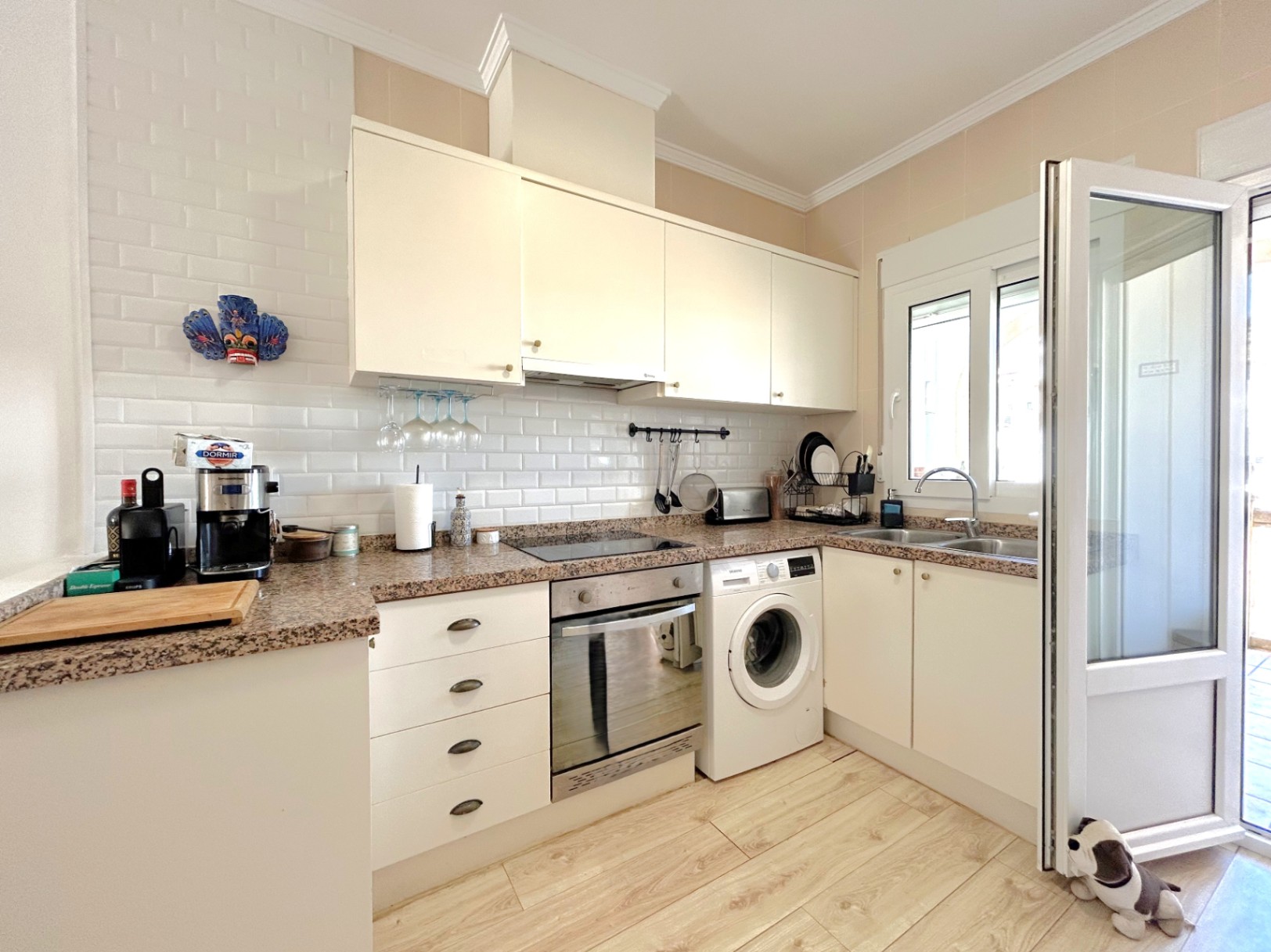 Cuisine équipée avec plan de travail en granit, meubles blancs et crédence en carrelage métro. Comprend des appareils intégrés et un sol en céramique effet bois.
