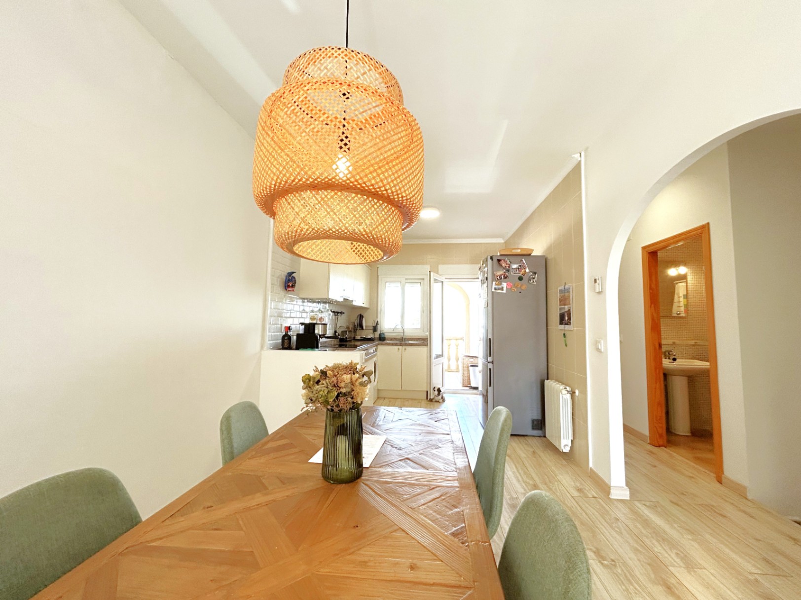 Espace salle à manger avec table en bois et suspension en rotin, ouvert sur une cuisine blanche équipée et un accès vers la buanderie lumineuse.