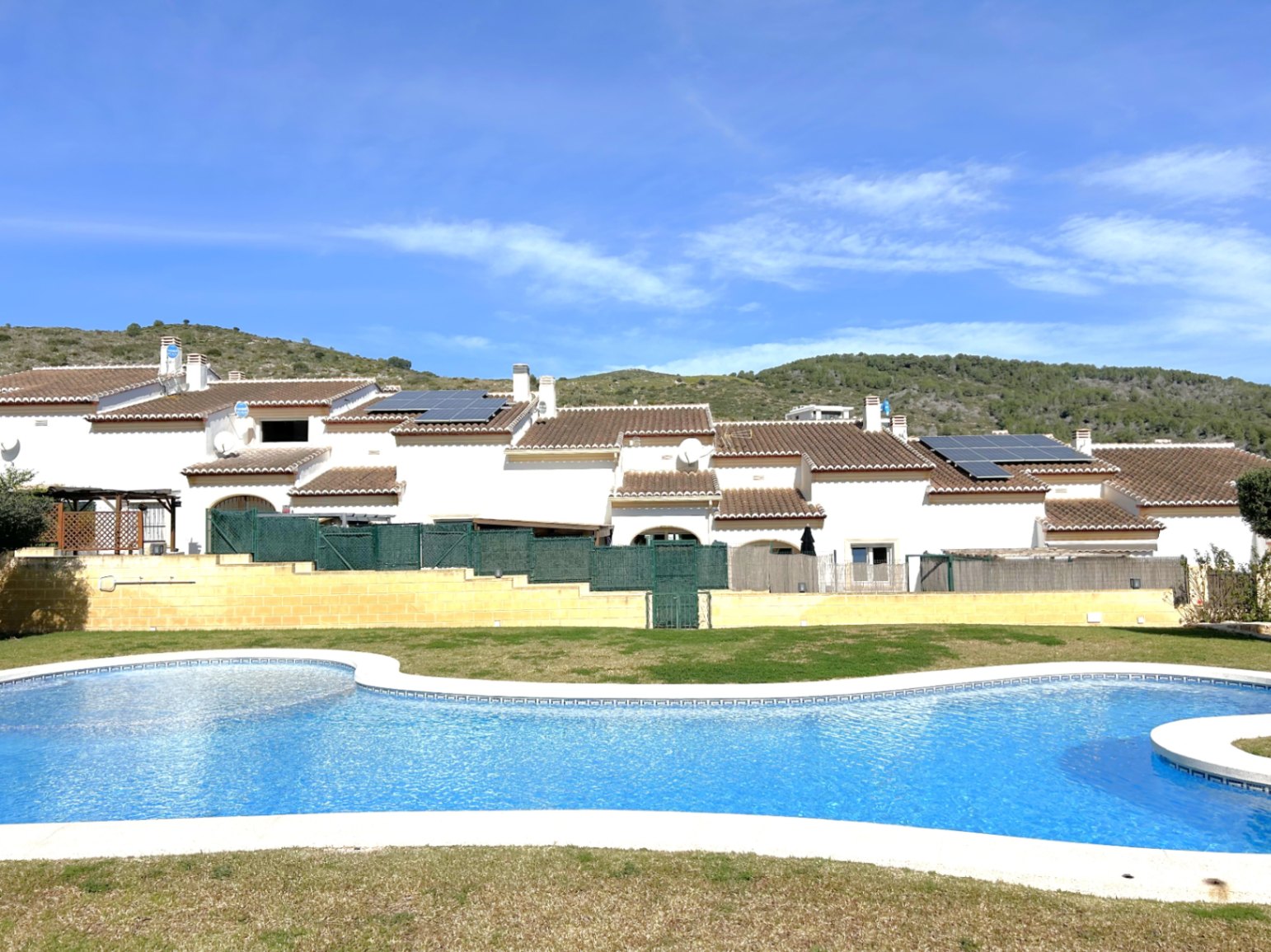 Grande piscine communautaire aux formes arrondies entourée de pelouse, avec en arrière-plan les maisons mitoyennes blanches et les collines environnantes.