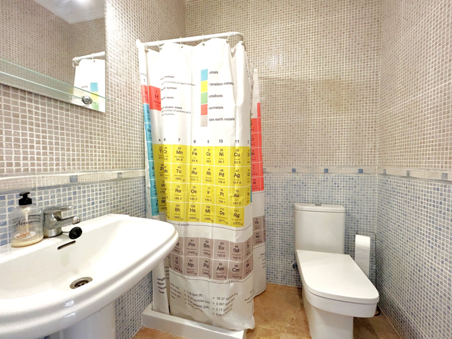 Salle de bain avec carrelage en mosaïque bicolore, lavabo sur colonne blanc et rideau de douche tableau périodique. Équipée d'un WC moderne et d'un miroir avec étagère en verre.
