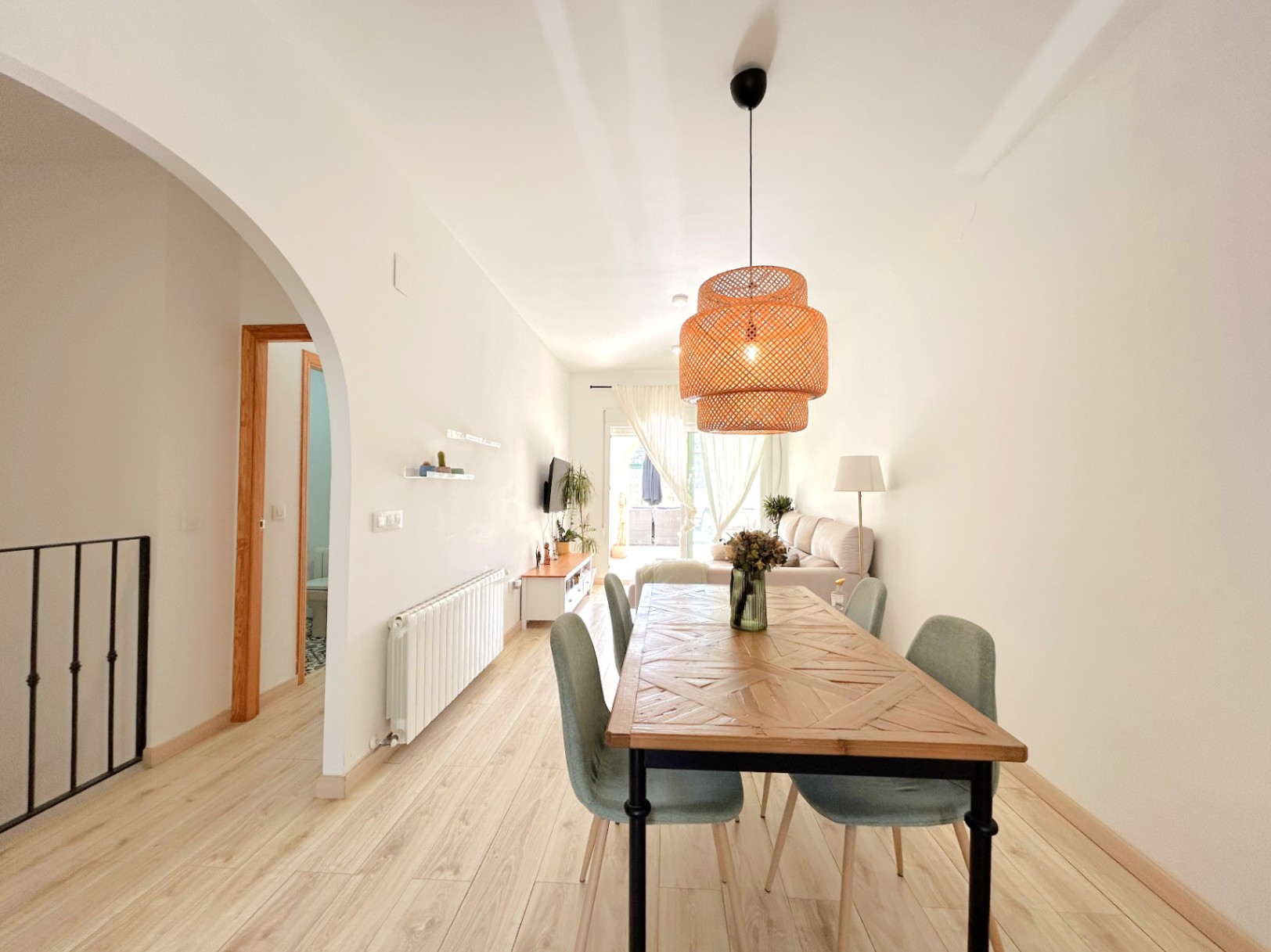 Séjour ouvert avec table à manger géométrique, luminaire en fibres naturelles et sol clair. L'espace dispose d'un radiateur mural et s'ouvre sur une terrasse lumineuse.