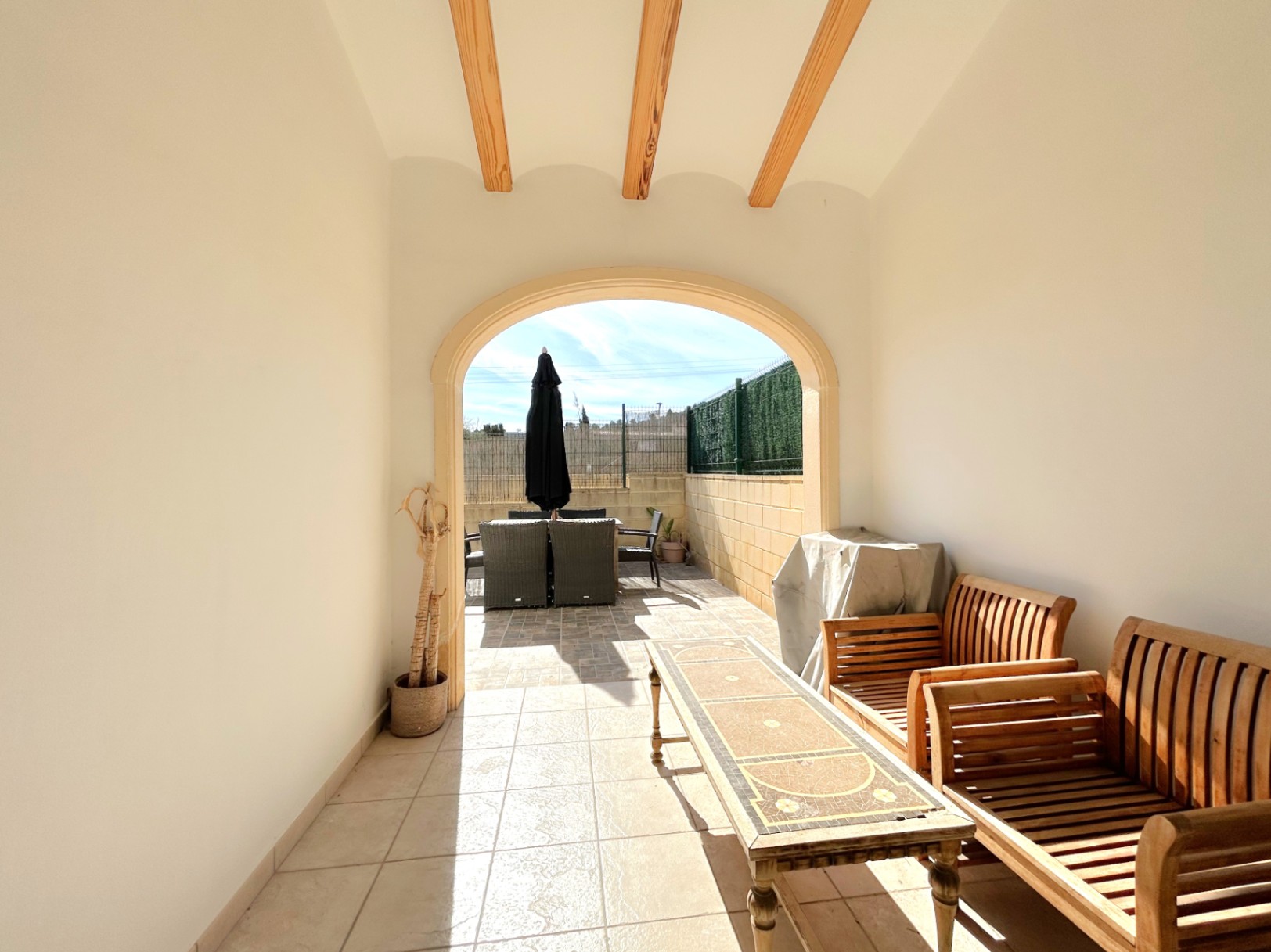 Terrasse couverte avec poutres en bois apparentes et arche élégante, offrant un espace de détente ombragé ouvert sur une terrasse ensoleillée.
