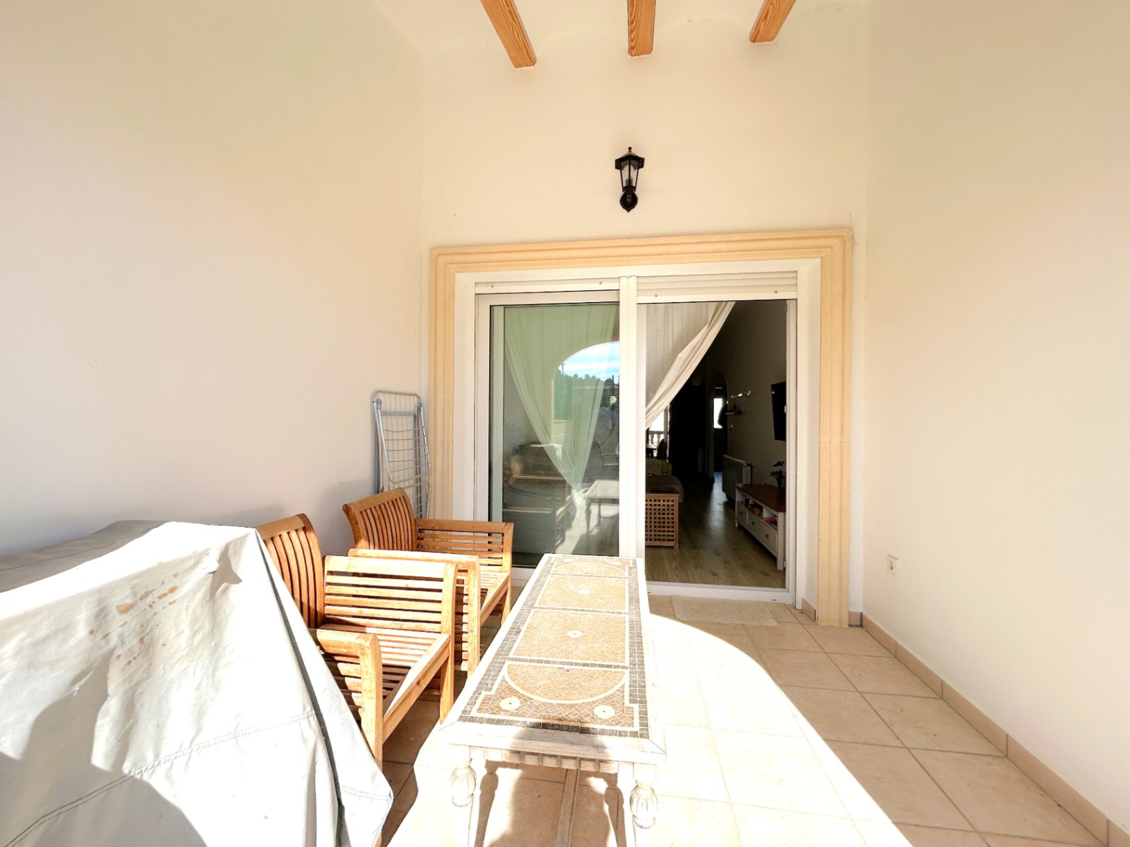 Terrasse couverte avec poutres en bois apparentes, sol en céramique et mobilier. Une grande baie vitrée relie l'extérieur au salon principal.